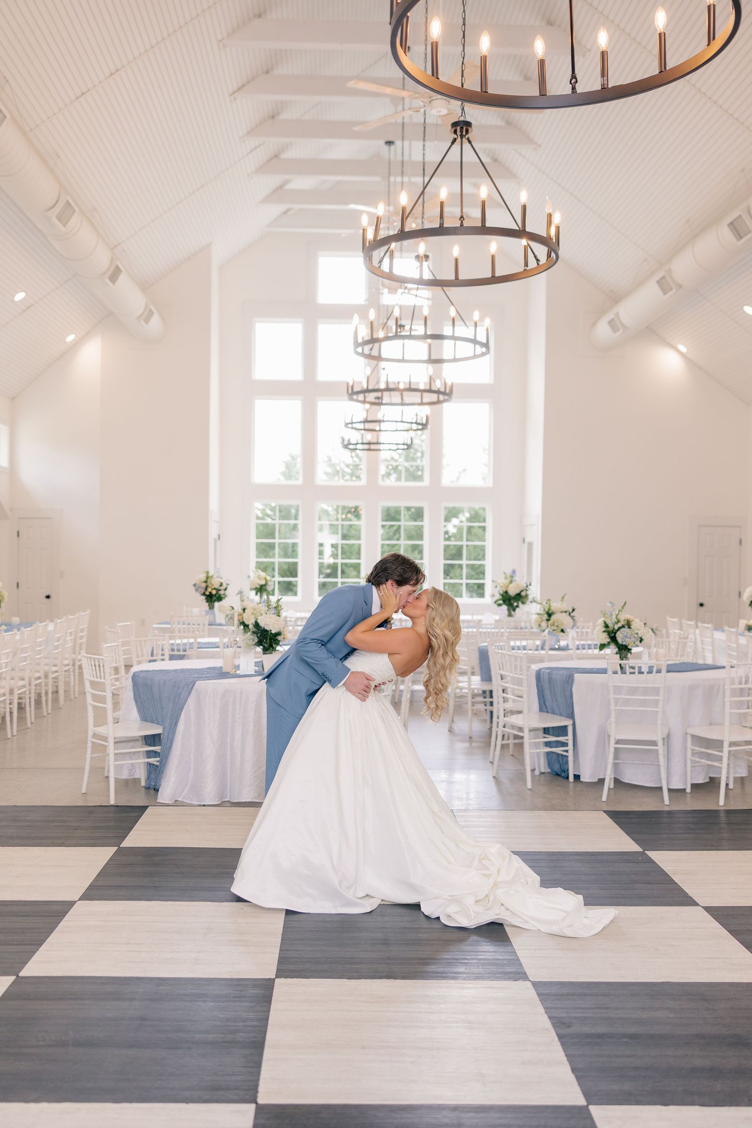 Bride and groom share a romantic dip kiss on a checkered dance floor in an elegant white reception hall.