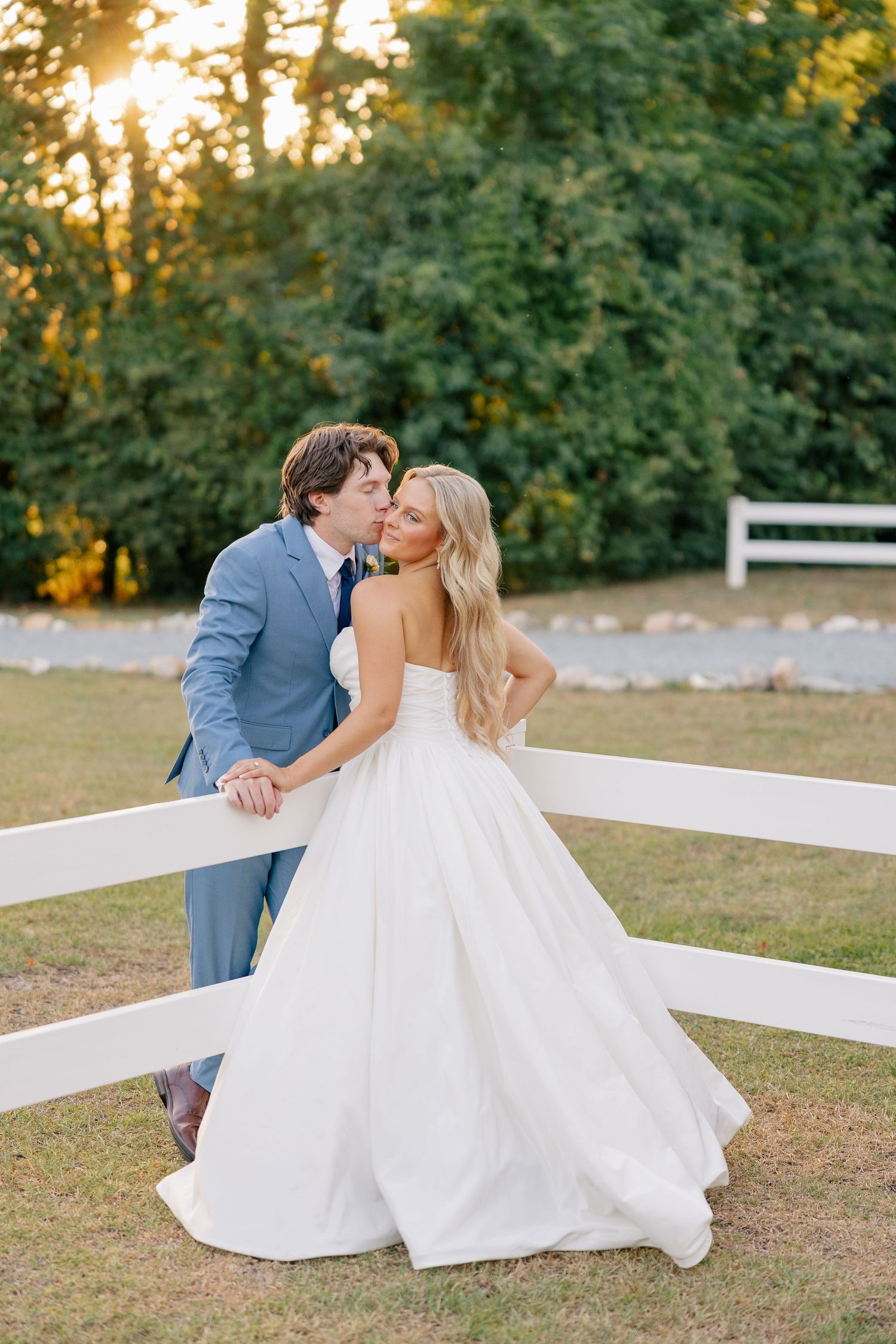 Bride and groom share a romantic kiss by a white fence at golden hour sunset.