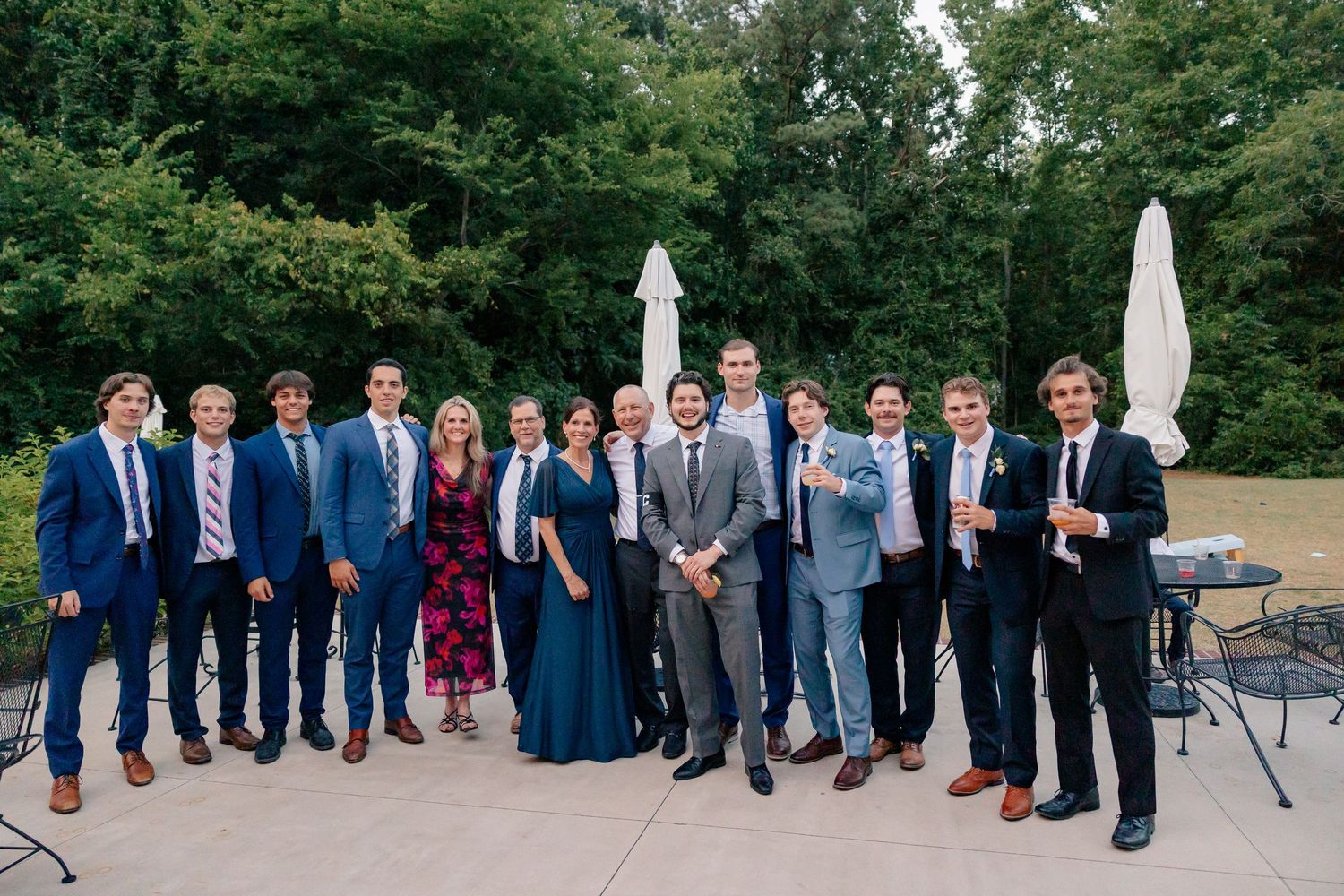 Large wedding guest group photo outdoors on a patio, guests dressed in formal attire with lush greenery behind.