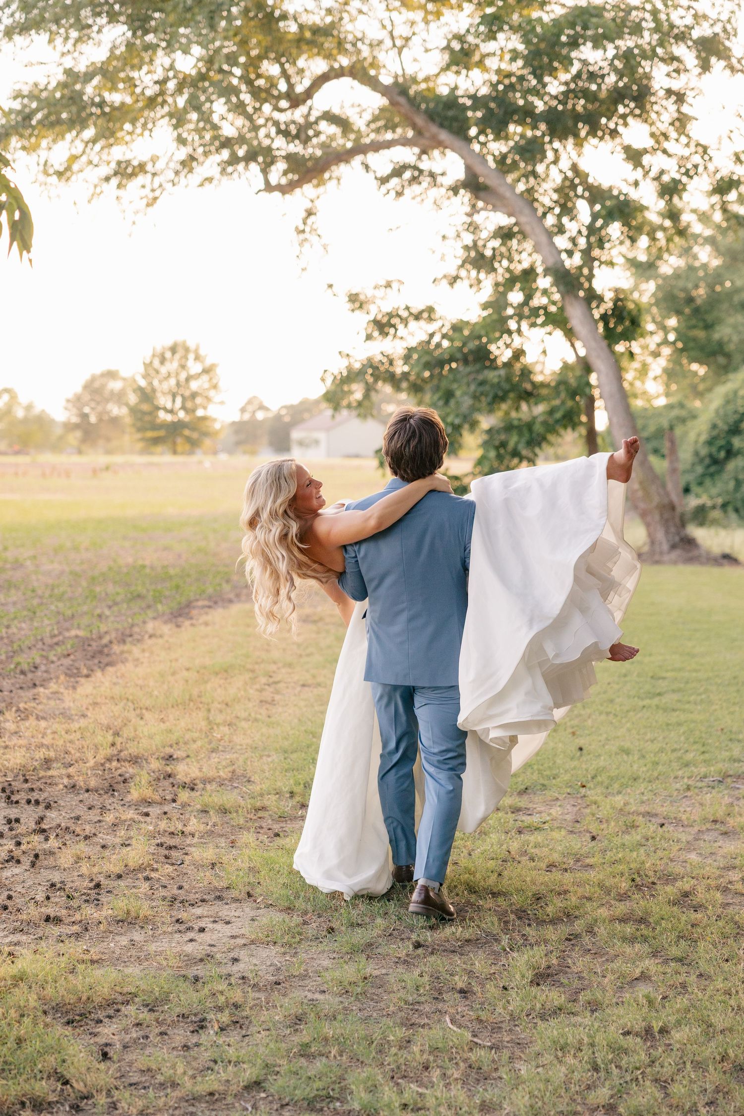 Groom carrying bride in white wedding gown across green field at golden hour.