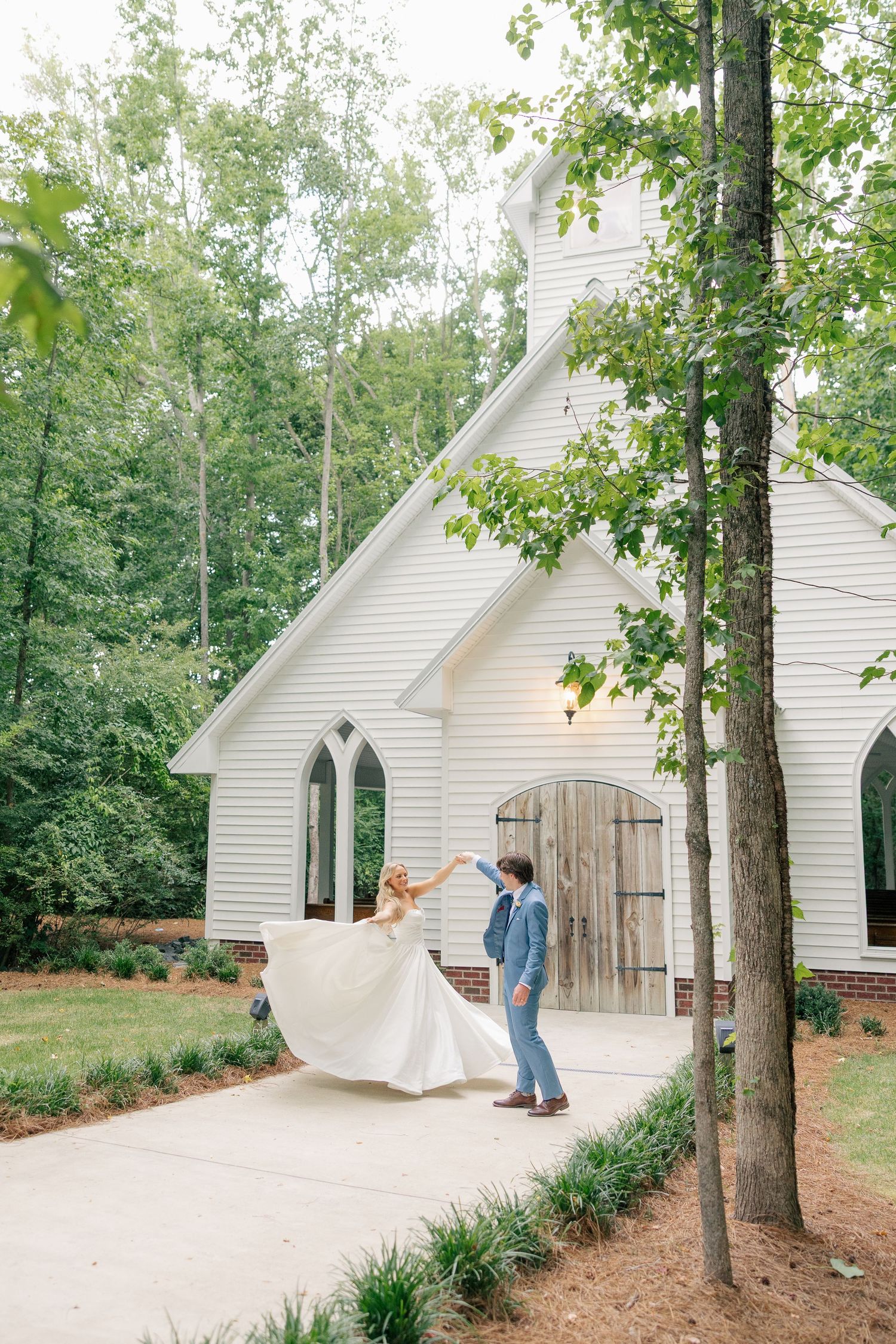 Groom twirls bride in white gown outside a charming white chapel surrounded by lush greenery.