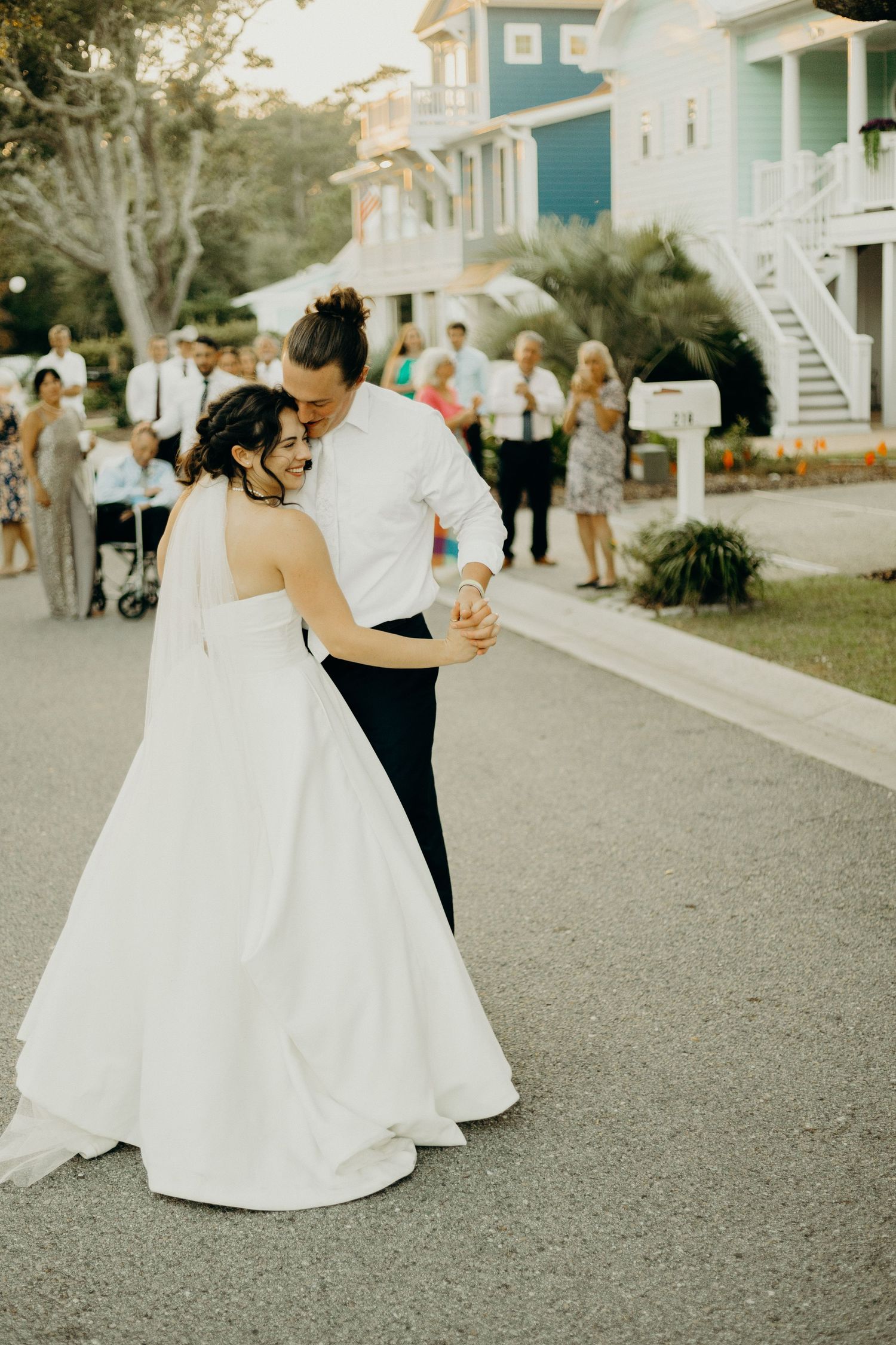 Newlywed couple shares an intimate dance on a paved path surrounded by white coastal homes and wedding guests.