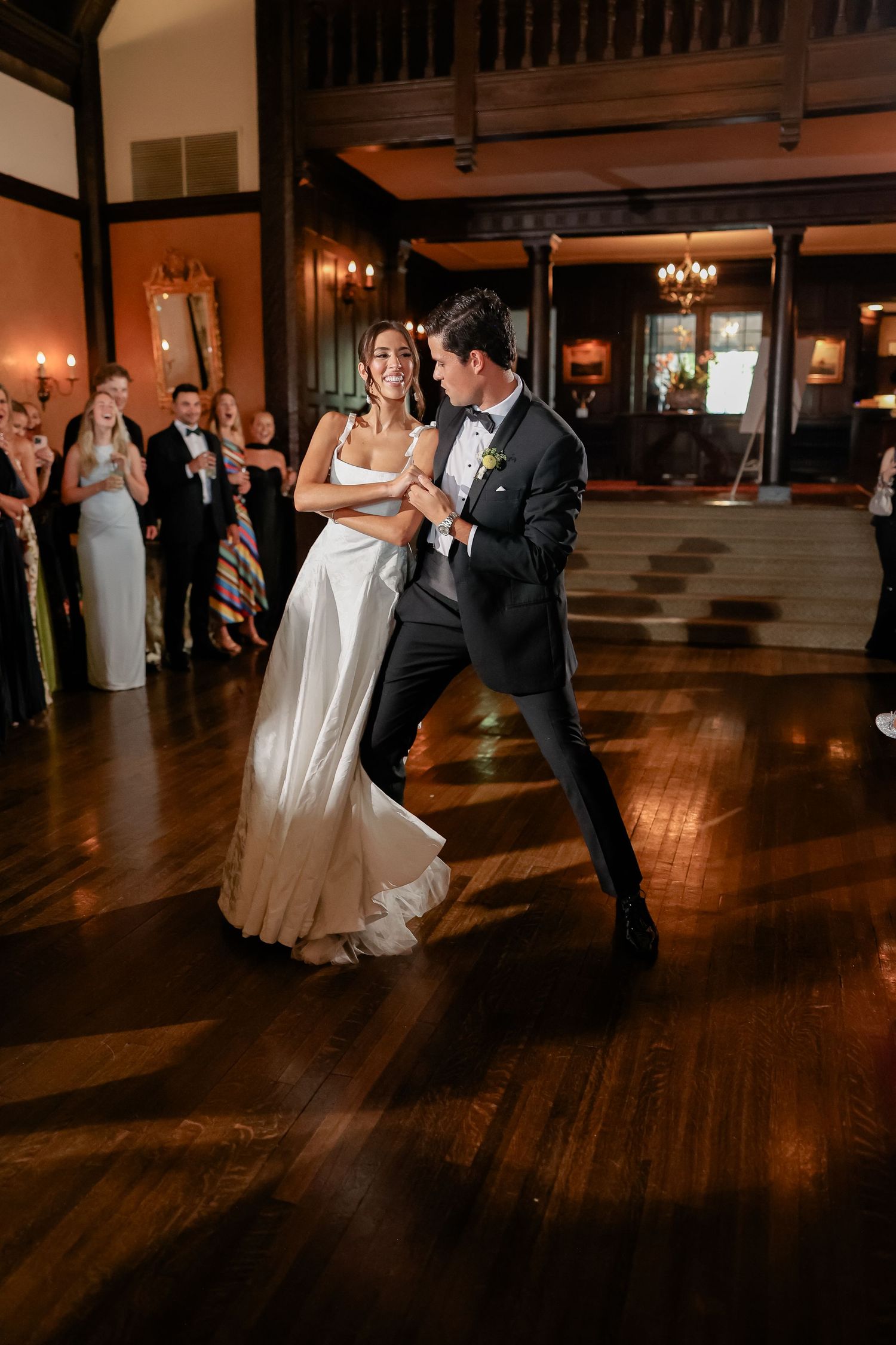Bride and groom share their first dance on a hardwood floor surrounded by applauding guests at a wedding reception.