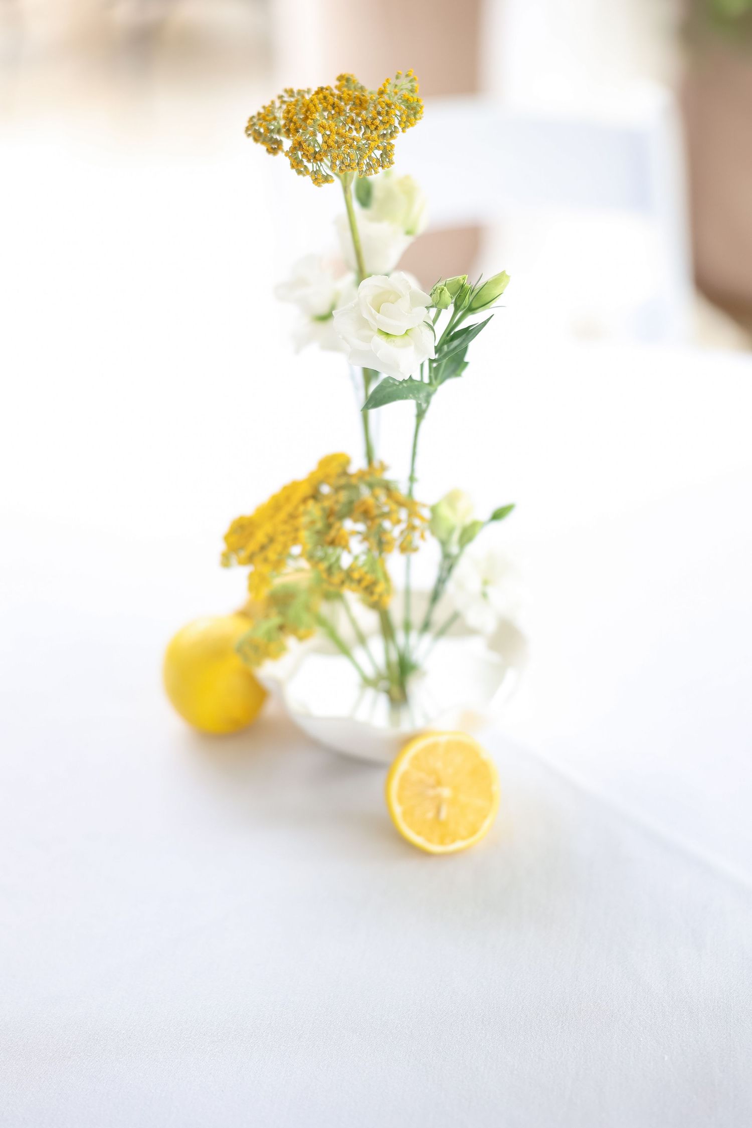 White vase with yellow wildflowers and white roses beside fresh lemons on a white tablecloth centerpiece.