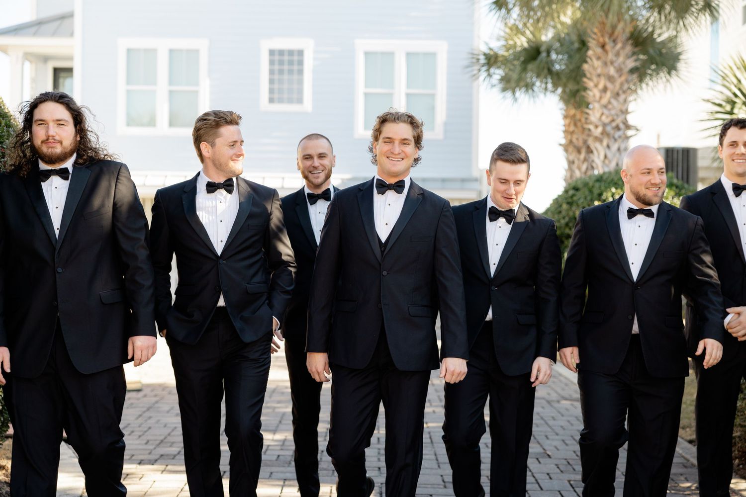 Wedding party of groomsmen walking and smiling together while wearing classic black tuxedos outdoors.