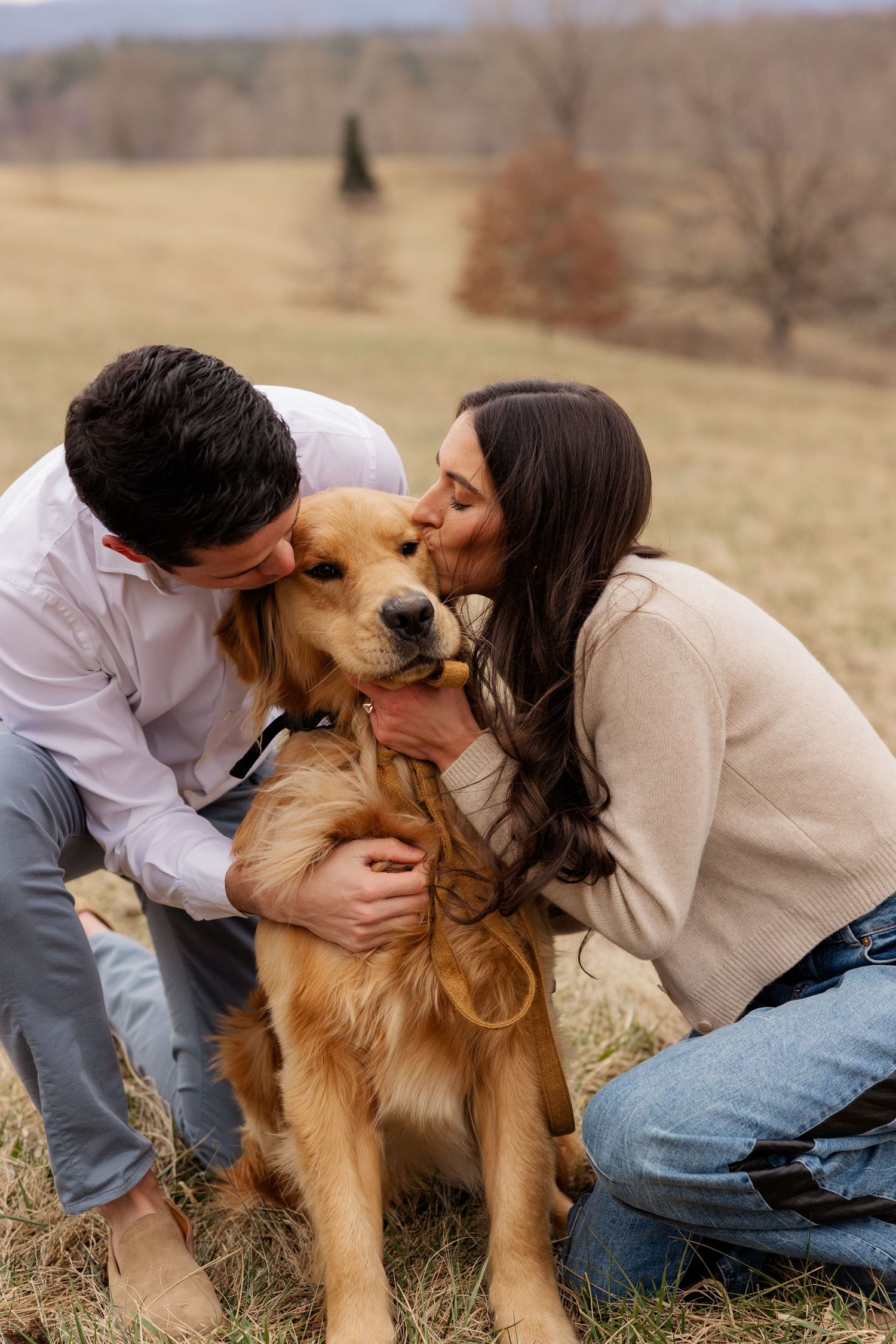 A couple shares sweet kisses with their happy golden retriever dog in a scenic autumn field.