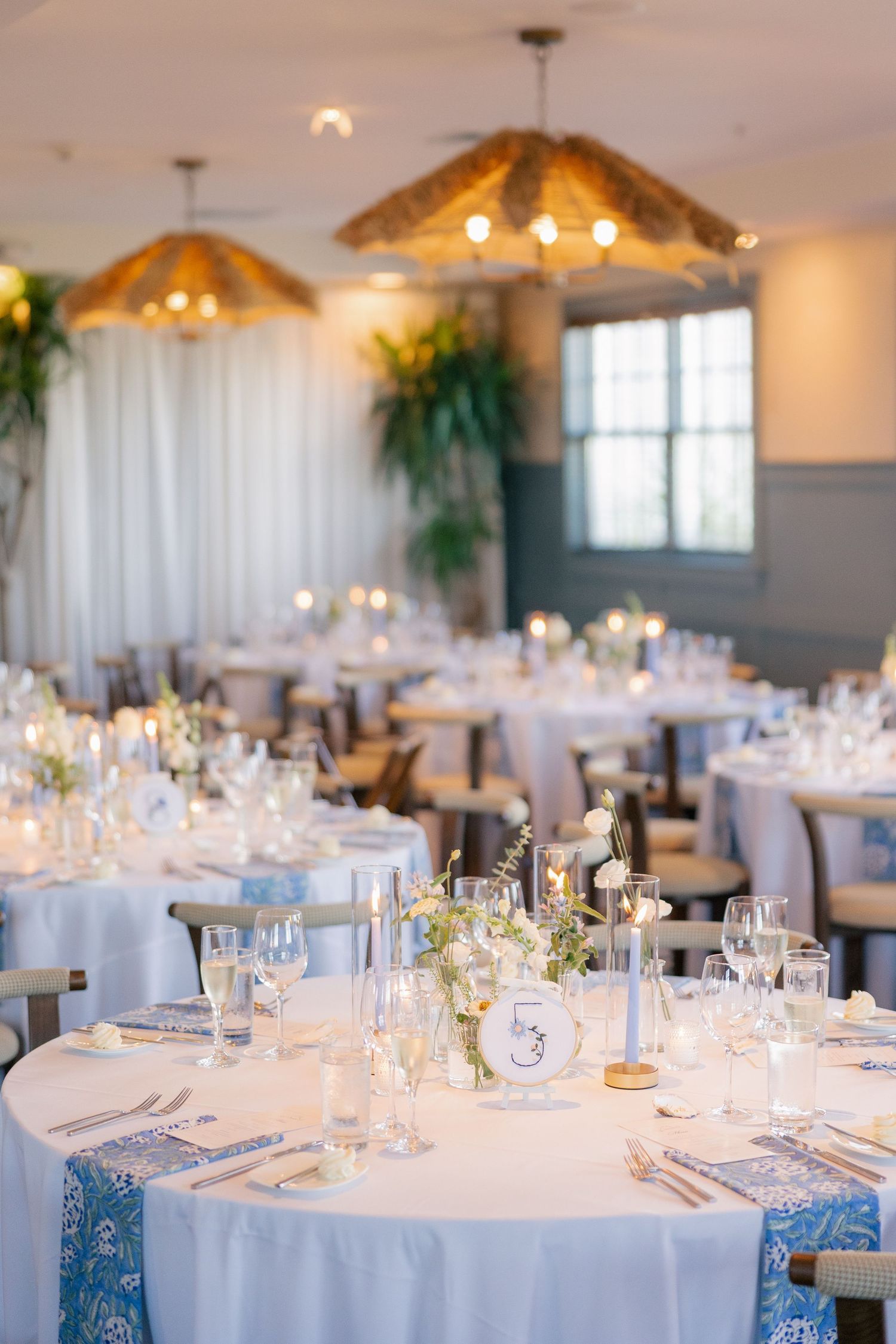 Elegant wedding reception hall with white round tables, blue accents, candles, and woven pendant lights.