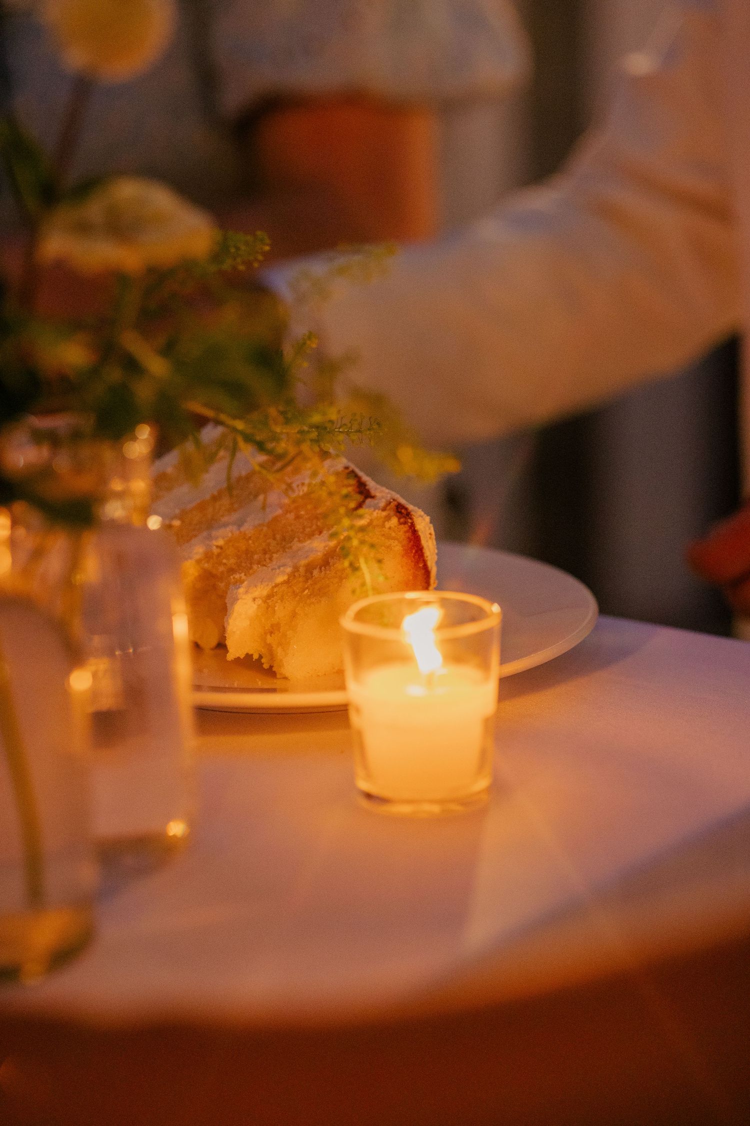 Slice of wedding cake on white plate beside a glowing candle and floral centerpiece at reception.