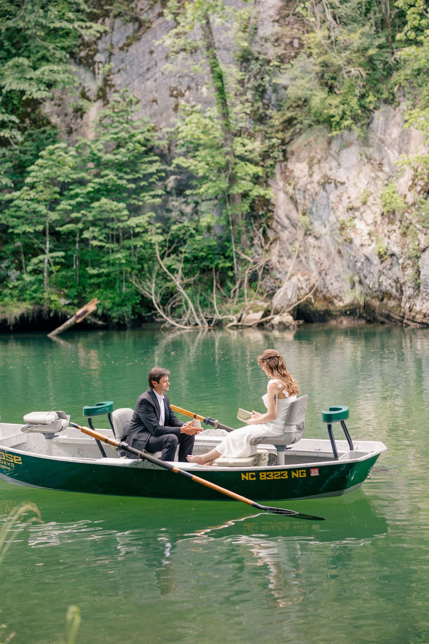 Couple in wedding attire seated in a green rowboat on a calm quarry lake.