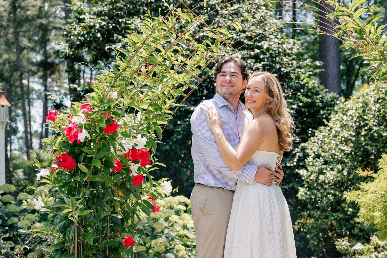 Couple embracing in a lush garden beside a blooming red rose bush, wearing elegant casual attire.