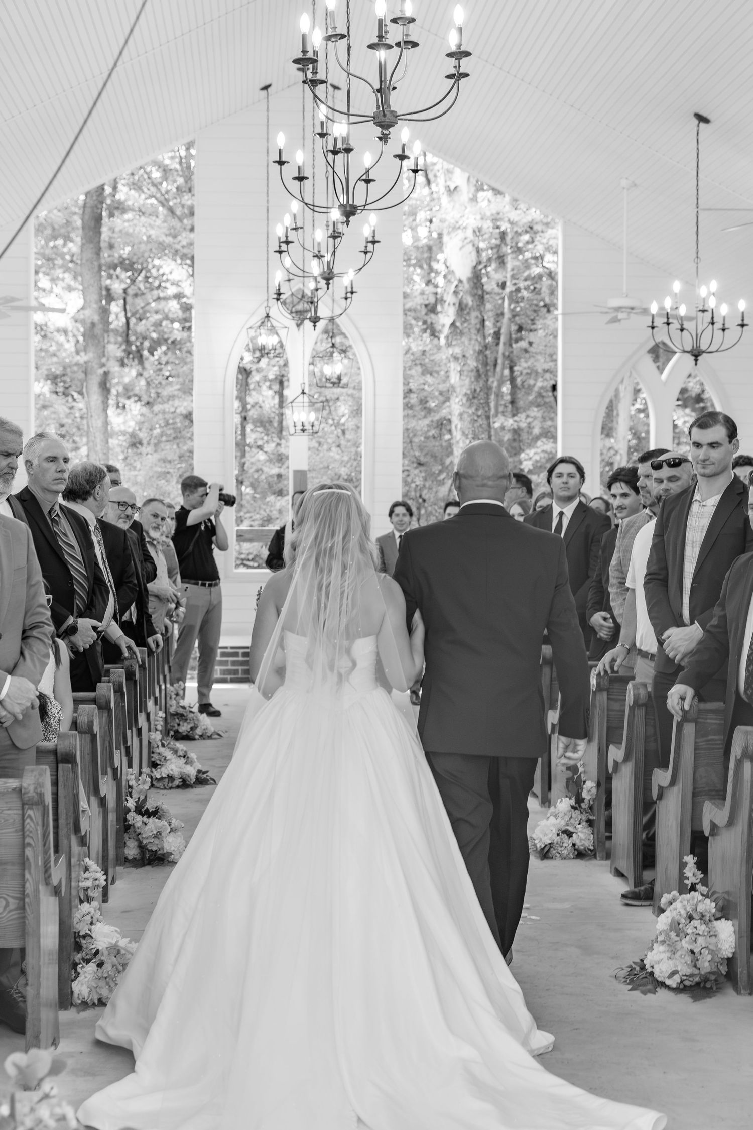 Bride walks down chapel aisle with escort in black and white wedding ceremony photo.