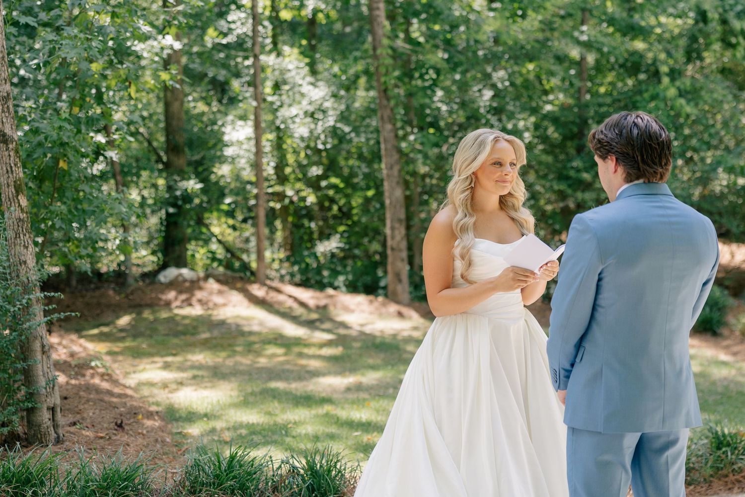 Bride in strapless white ballgown reads vows to groom in a lush green outdoor garden setting.