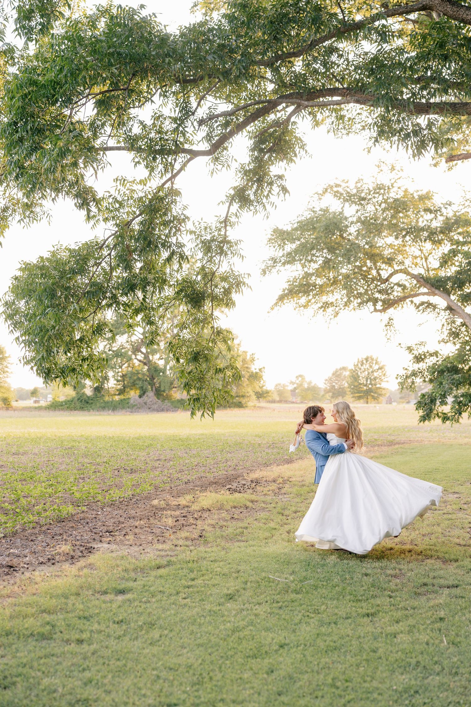 Groom lifting bride in white gown under a large oak tree at golden hour in an open field.