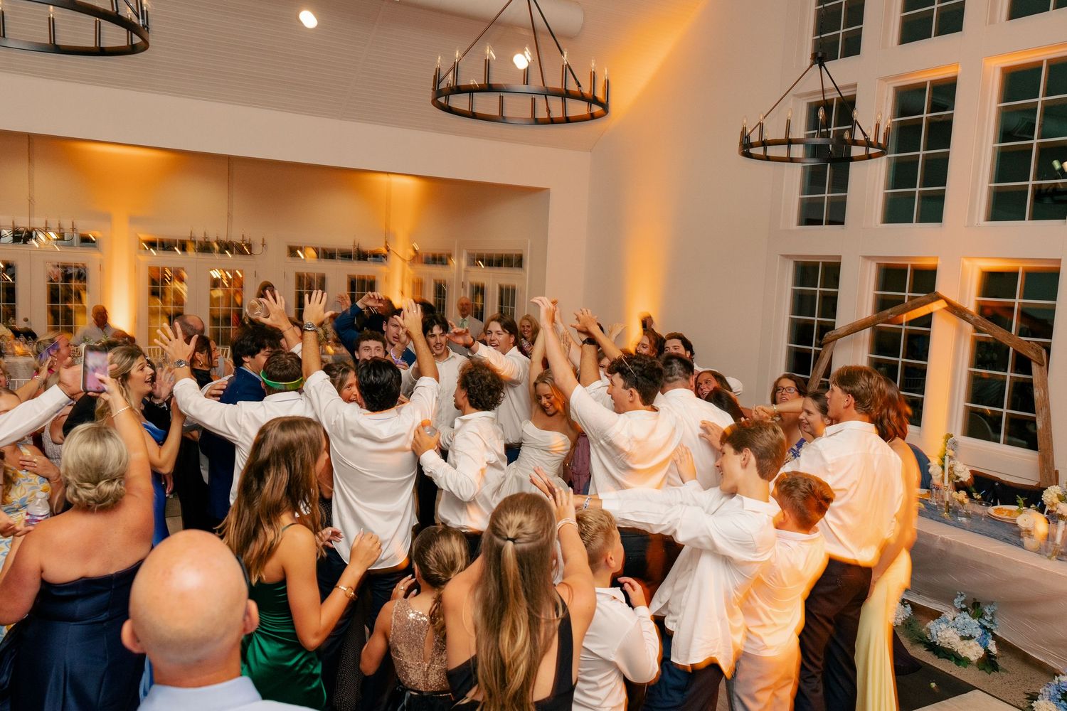 Wedding guests and bridal party celebrate on the dance floor with hands raised at a lively indoor reception.