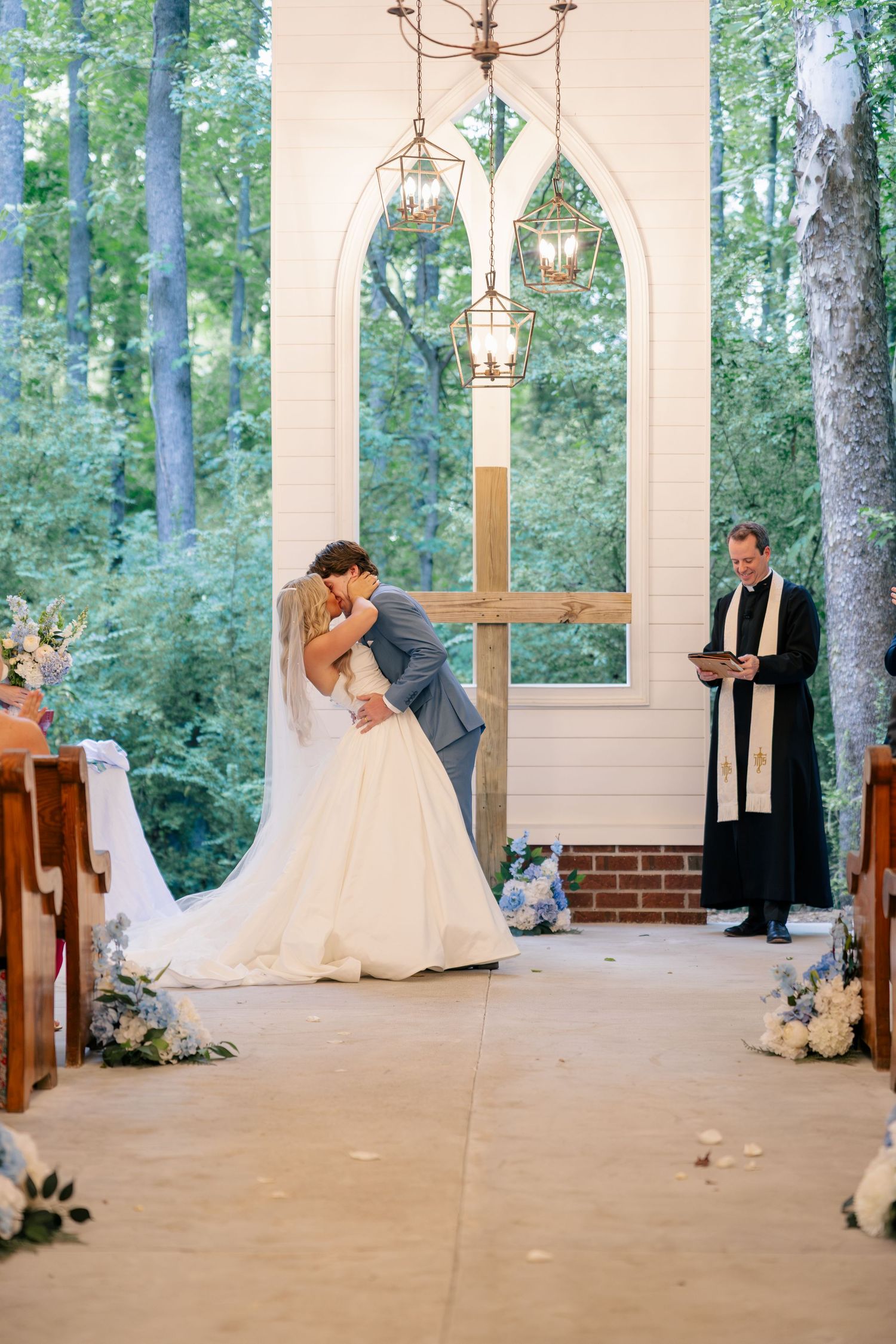 Bride and groom sharing first kiss at altar in white chapel with wooden cross and officiant.