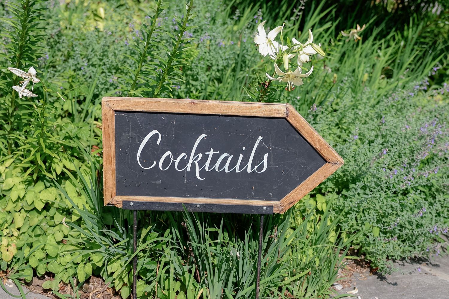 Wooden arrow-shaped chalkboard sign with 'Cocktails' written in white lettering among garden flowers.