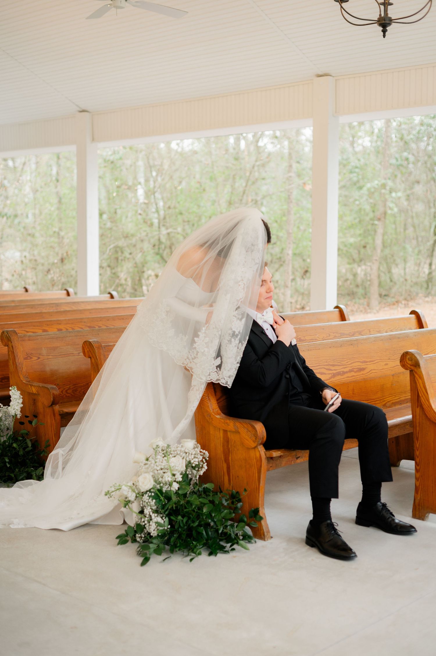 Wedding couple playfully hiding behind wooden church pews decorated with greenery.
