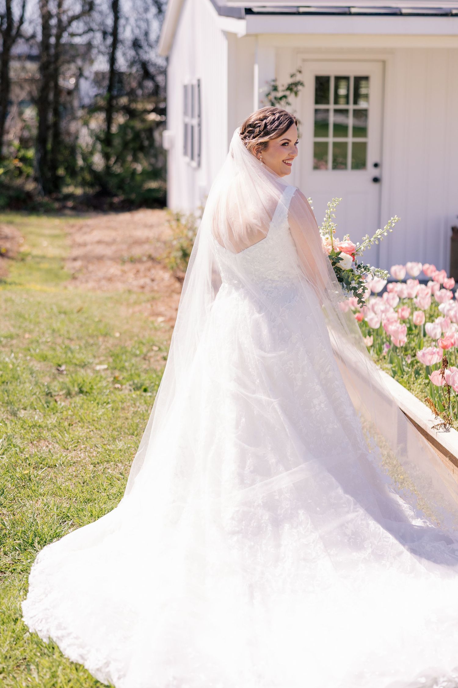 A flowing white wedding dress and veil photographed outdoors against a white building and garden setting.