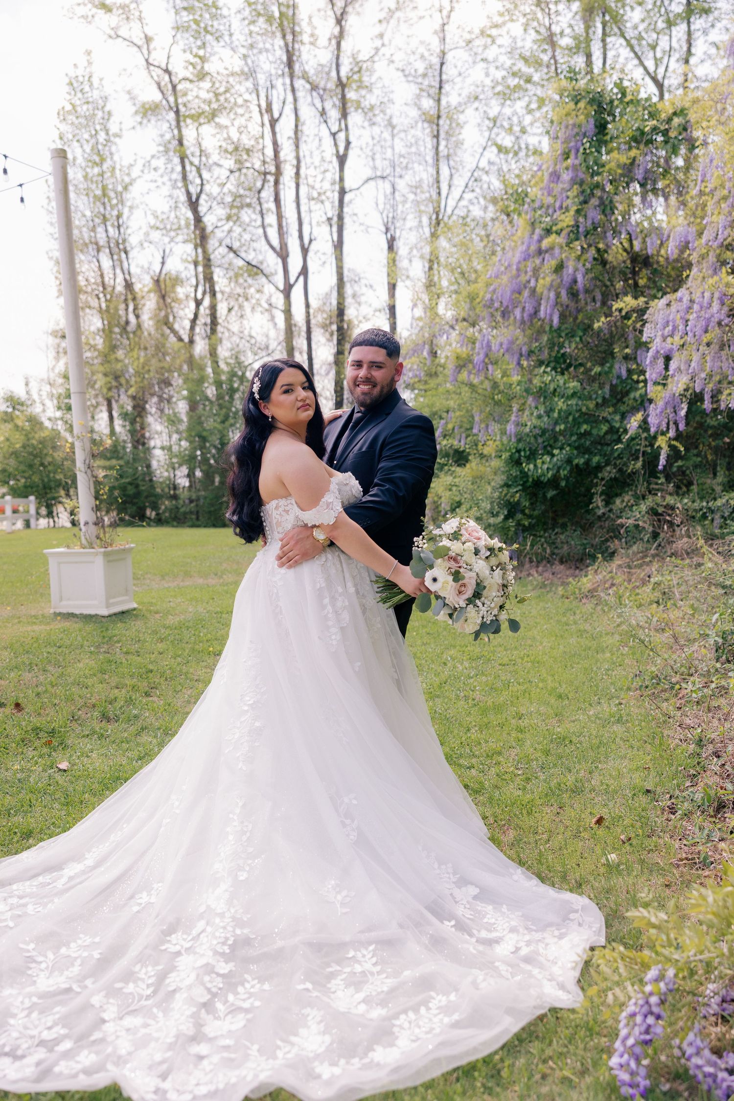 Wedding couple poses in a garden setting with blooming purple wisteria and a flowing lace wedding gown.