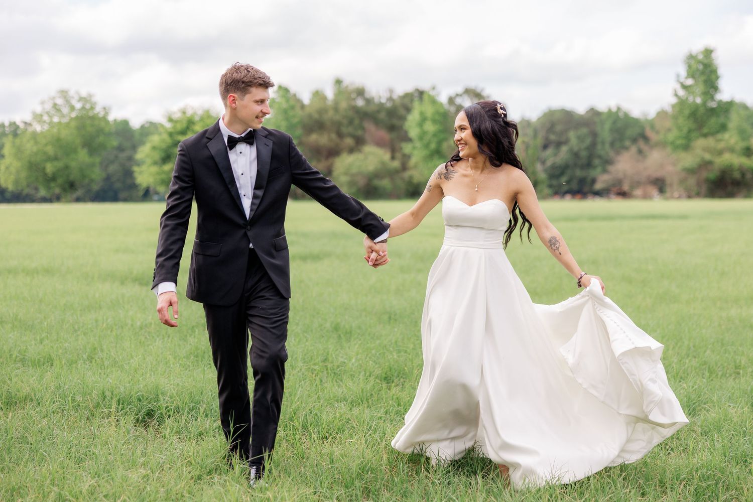 A couple in wedding attire walking hand in hand across a green field.