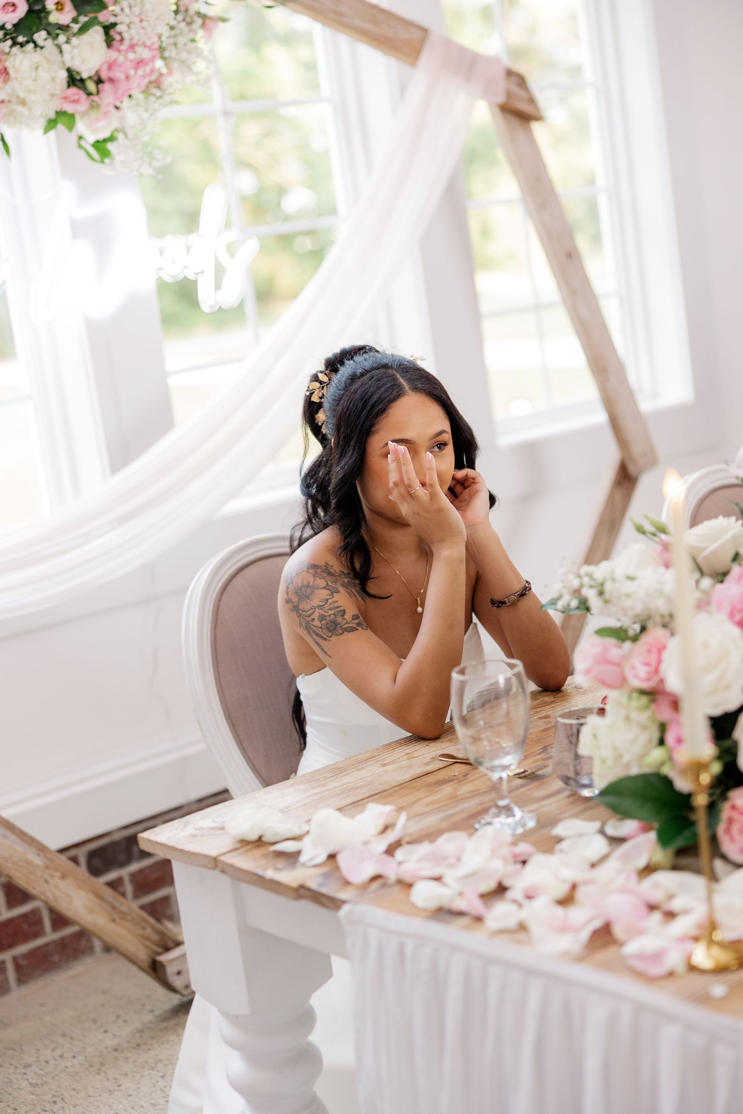 A bride in white dress sits at decorated table with floral arrangements.
