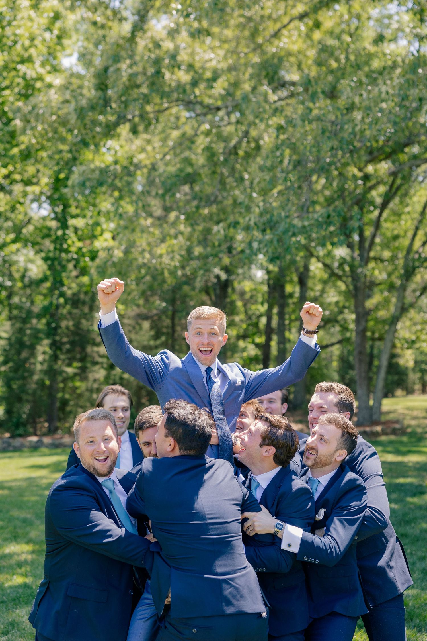 Groomsmen in navy suits lift celebrating groom with raised fists in sunny outdoor wedding party photo.