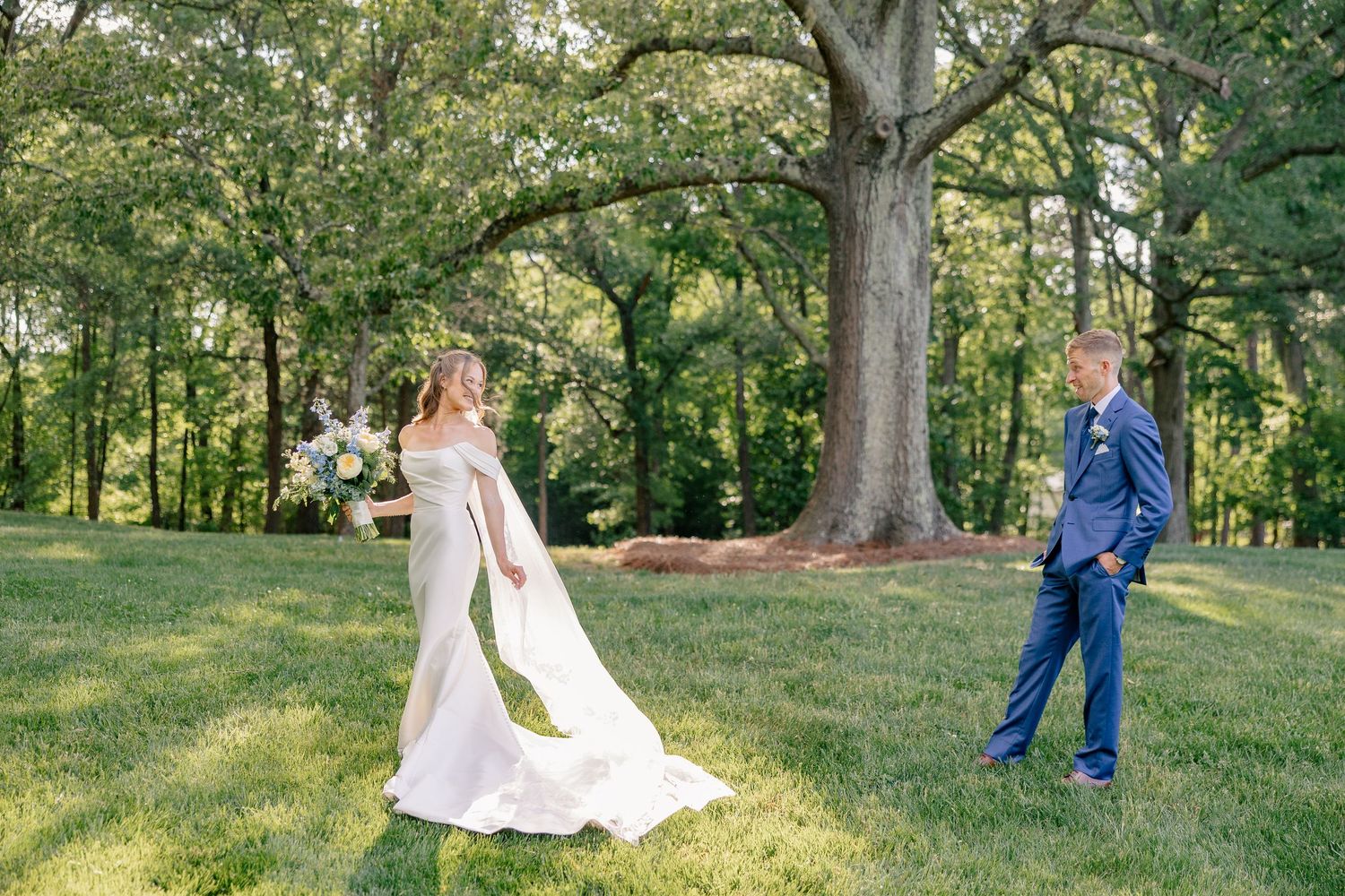 Bride in off-shoulder gown twirling her train during first look with groom under large oak tree.