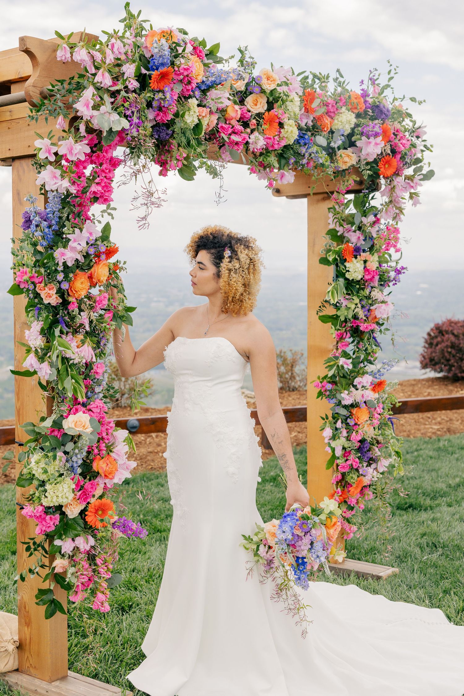Bride in white strapless gown stands under a vibrant rainbow floral wedding arch with mountain backdrop.