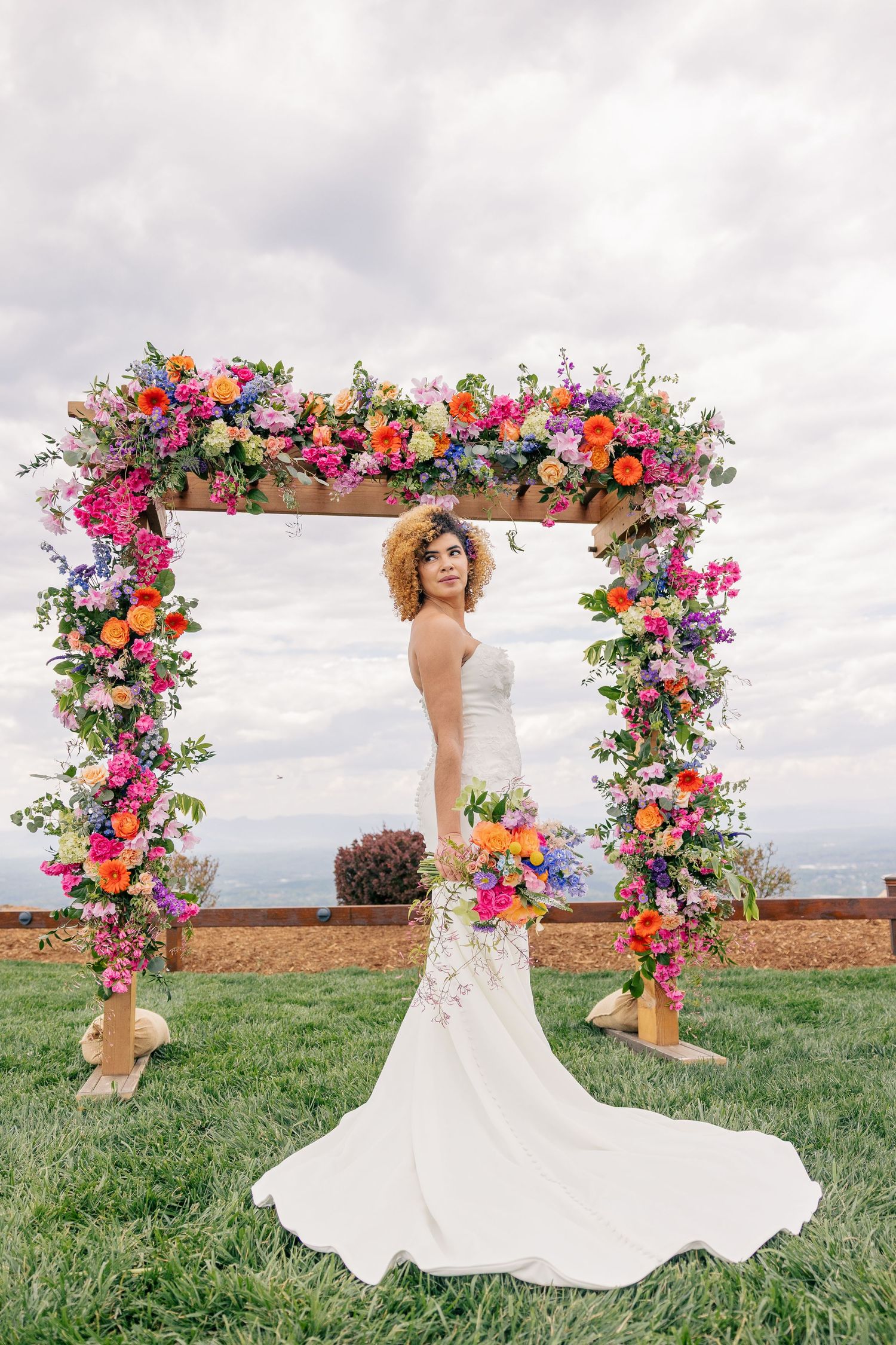 Bride in white gown poses under colorful floral arch with vibrant pink, orange, and purple blooms outdoors.