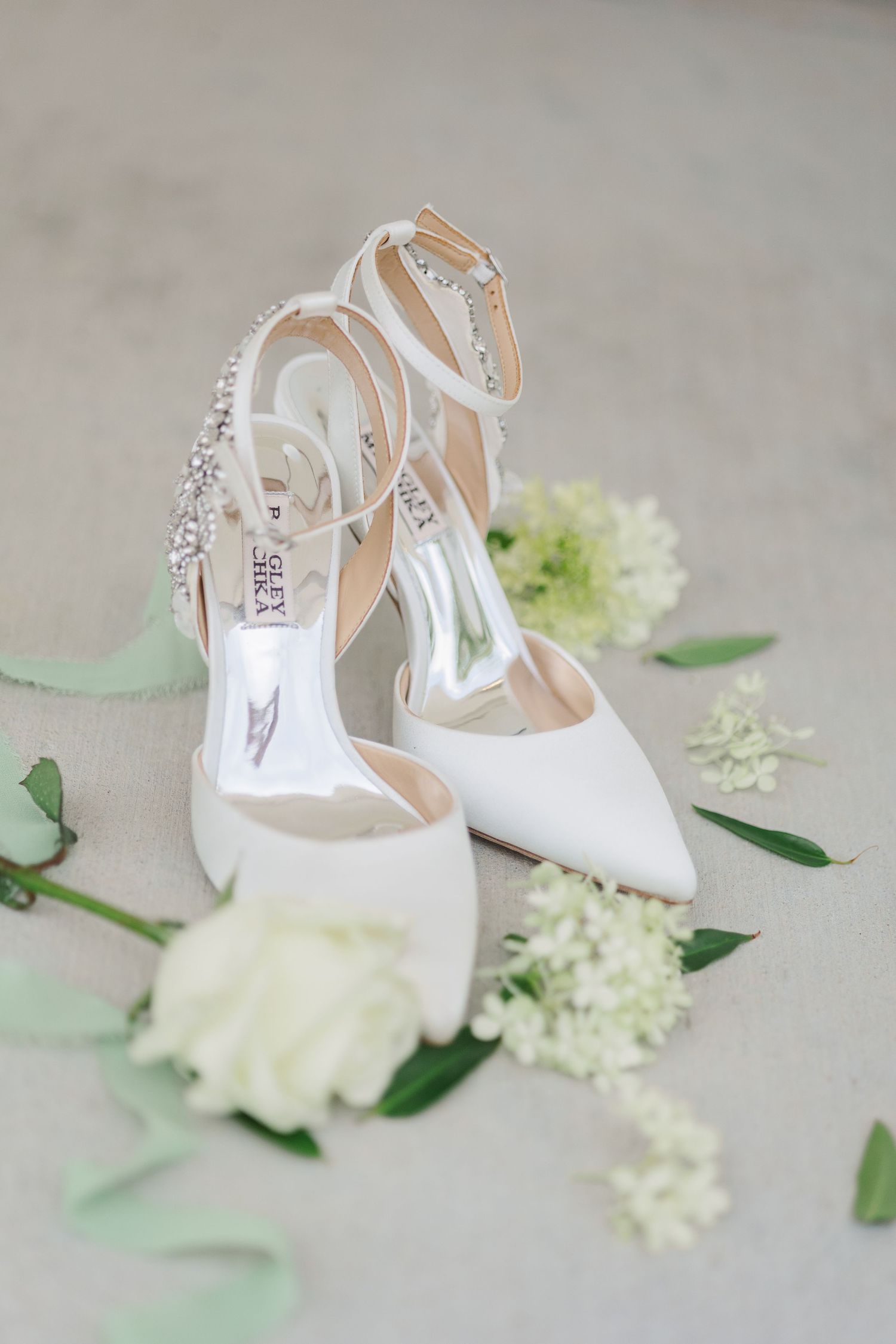 Elegant white bridal slingback heels displayed with delicate white flowers and green foliage on a light gray surface. Cornealius Properties