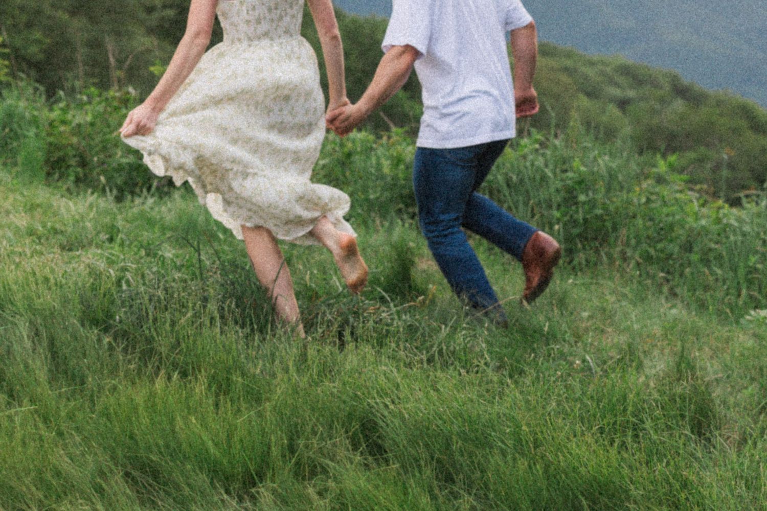A couple runs joyfully through a grassy hillside, with a flowing white dress catching the breeze.
