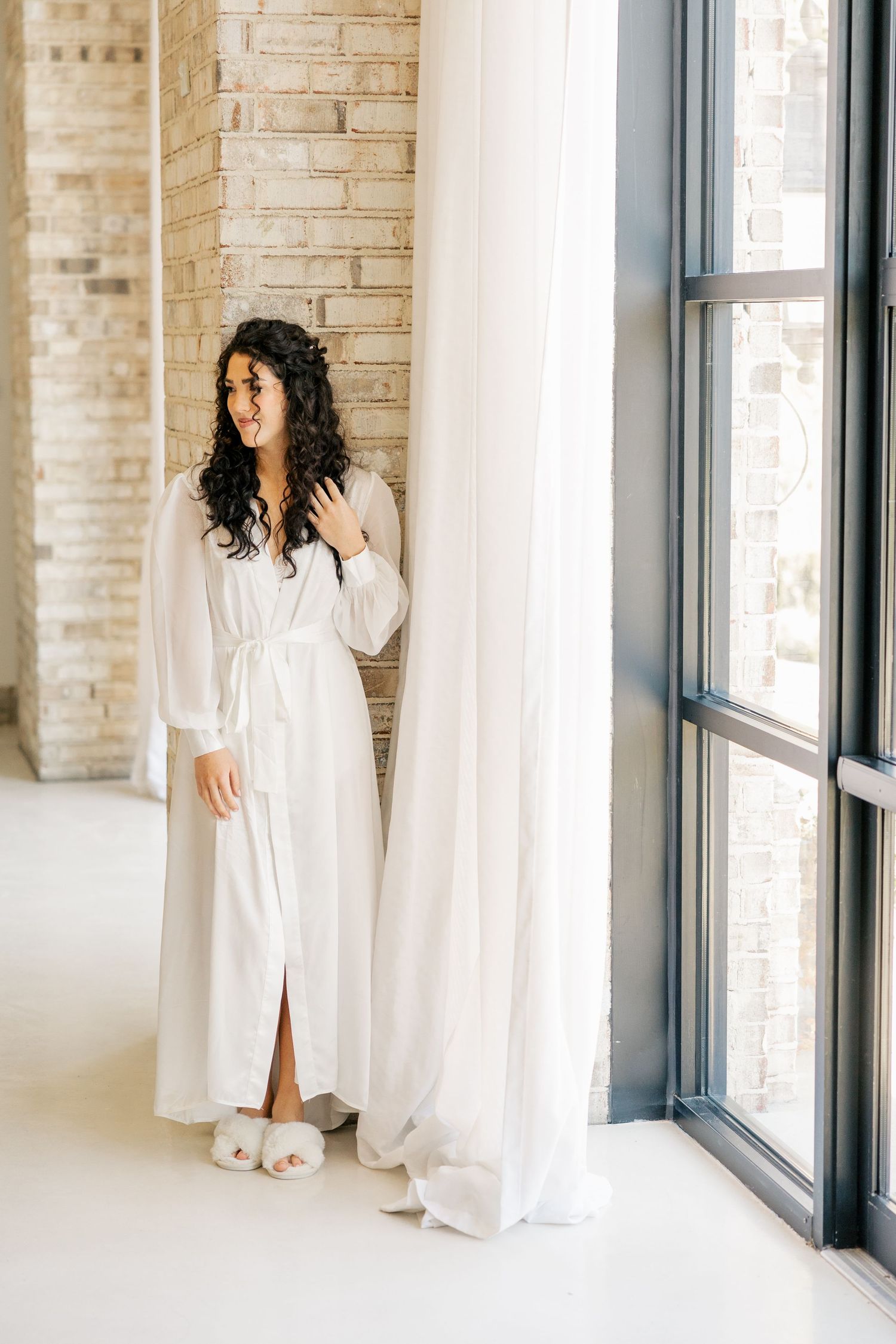 Woman in white robe standing near window with soft natural light and brick wall.