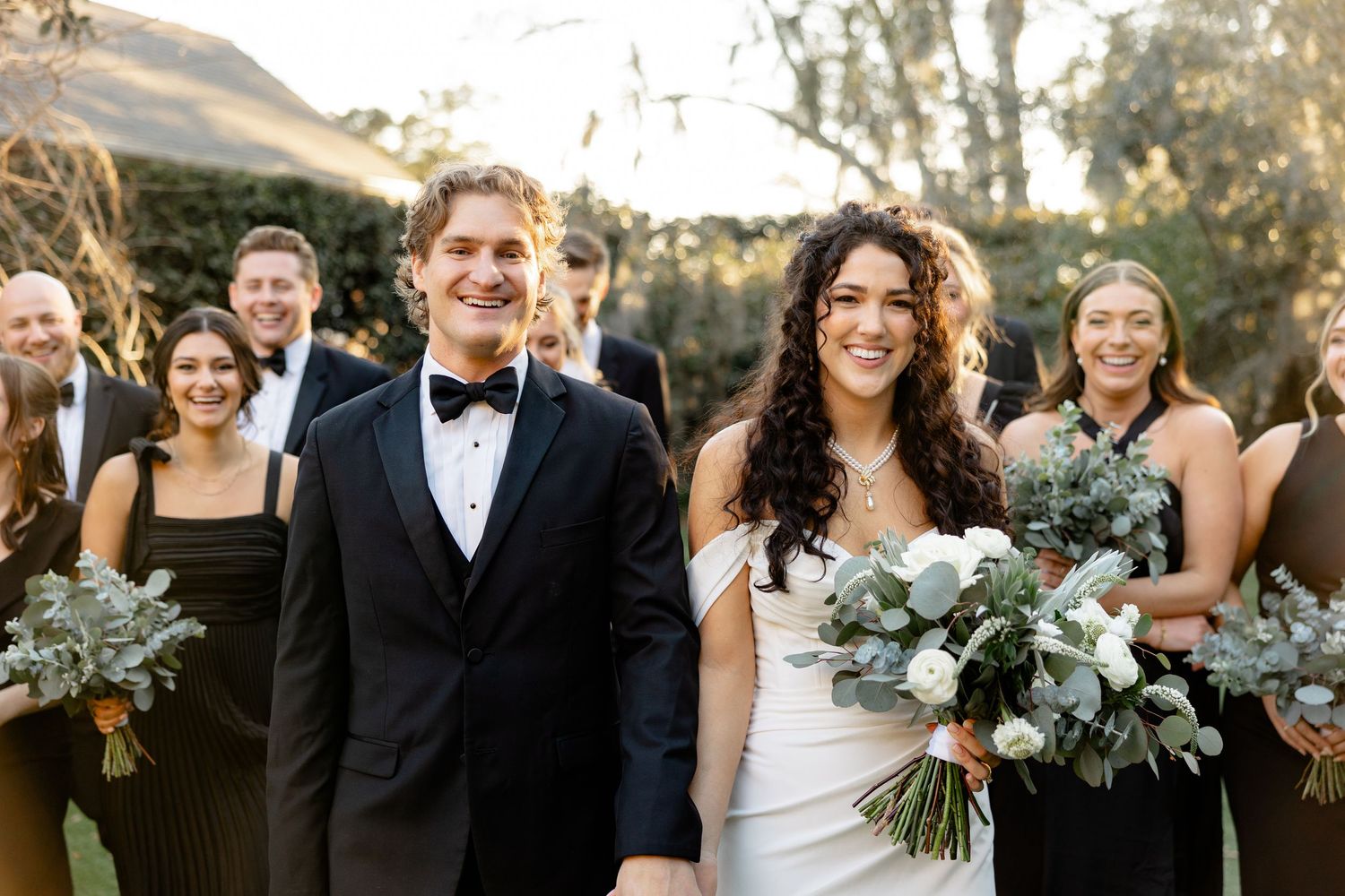 Happy newlyweds walking together outdoors with wedding party in background.