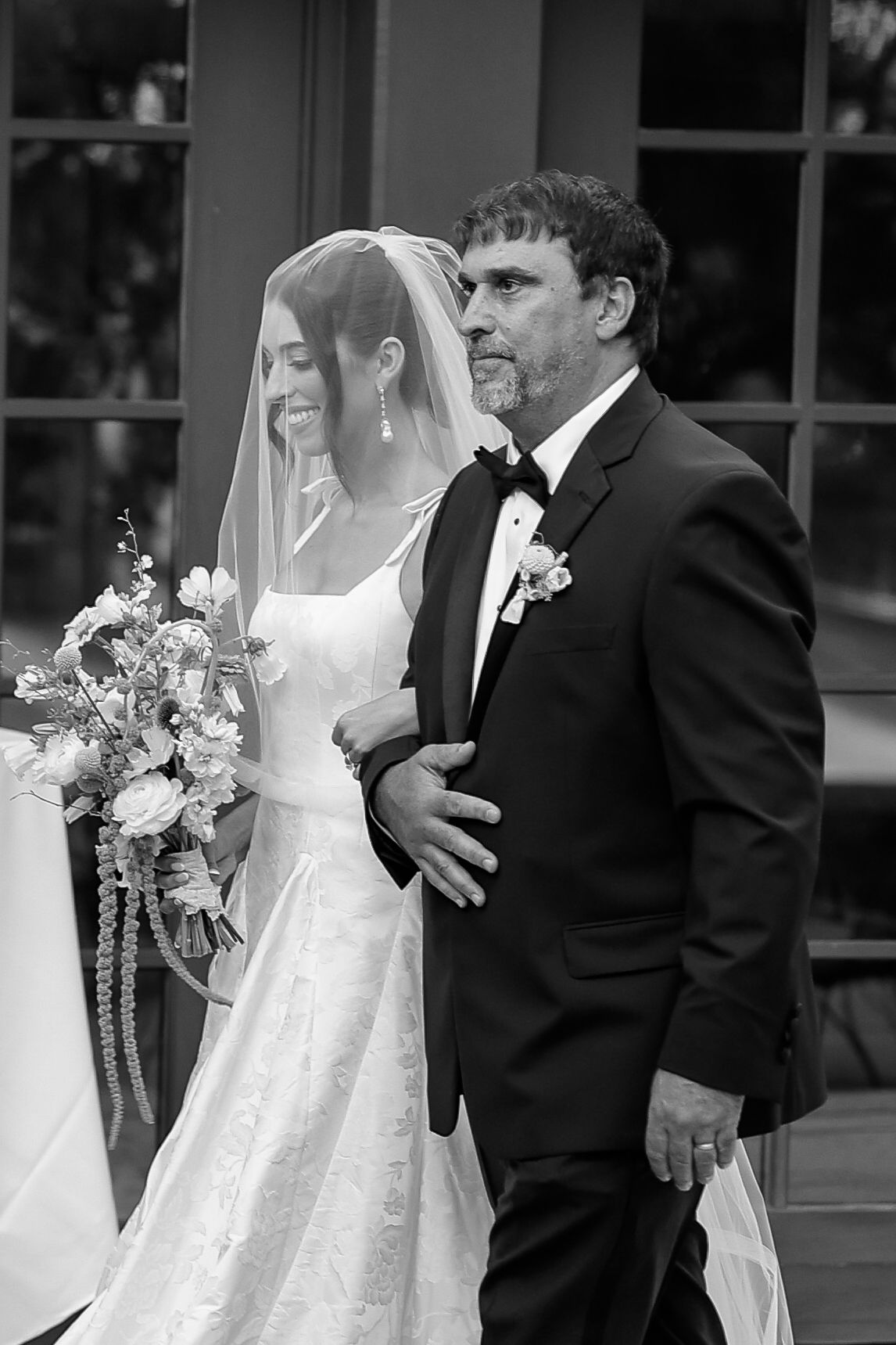 Black and white photo of a smiling bride in veil holding bouquet, escorted by a man in tuxedo at a wedding ceremony.