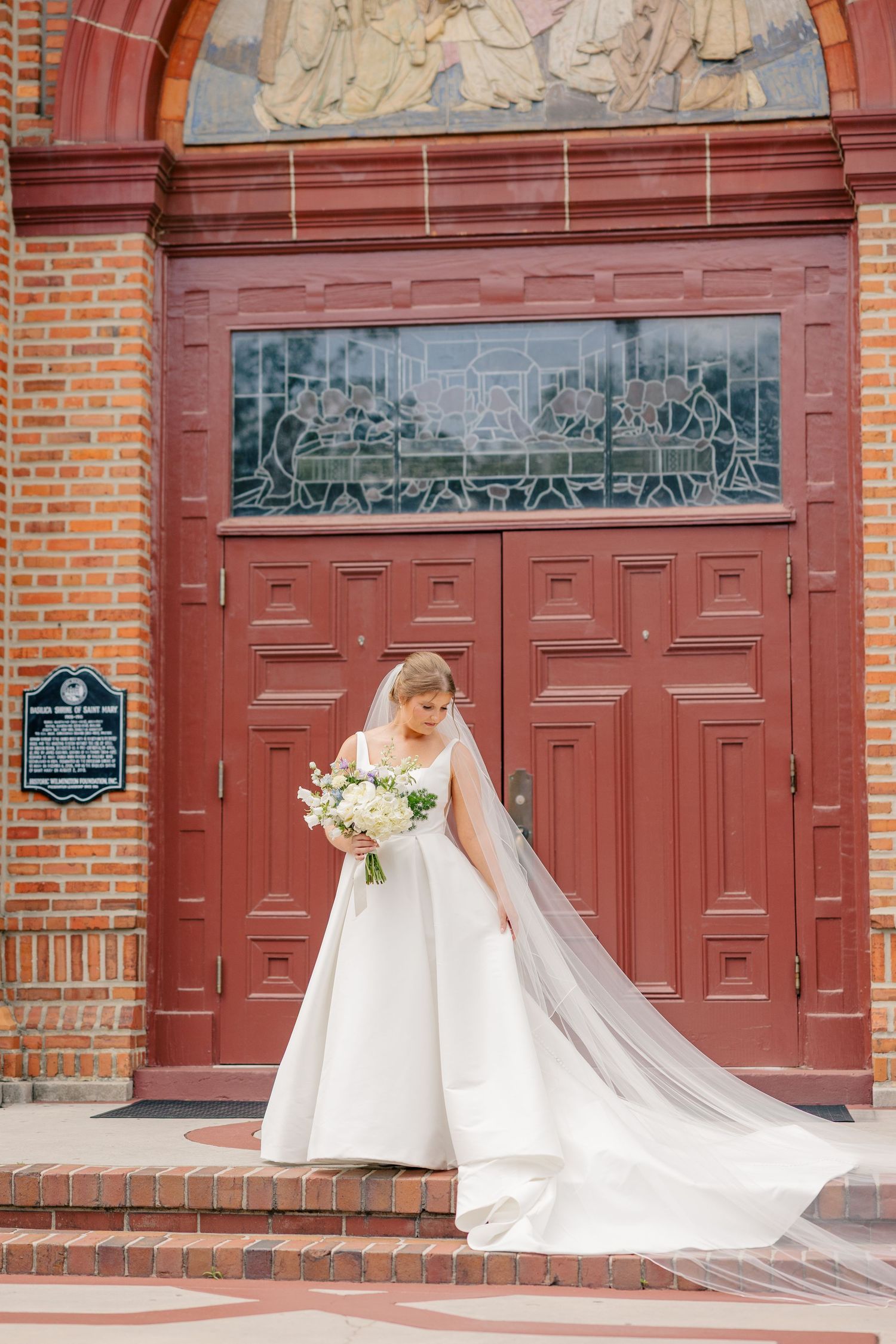 Bride in white ball gown with flowing veil stands before ornate red church doors holding white floral bouquet.