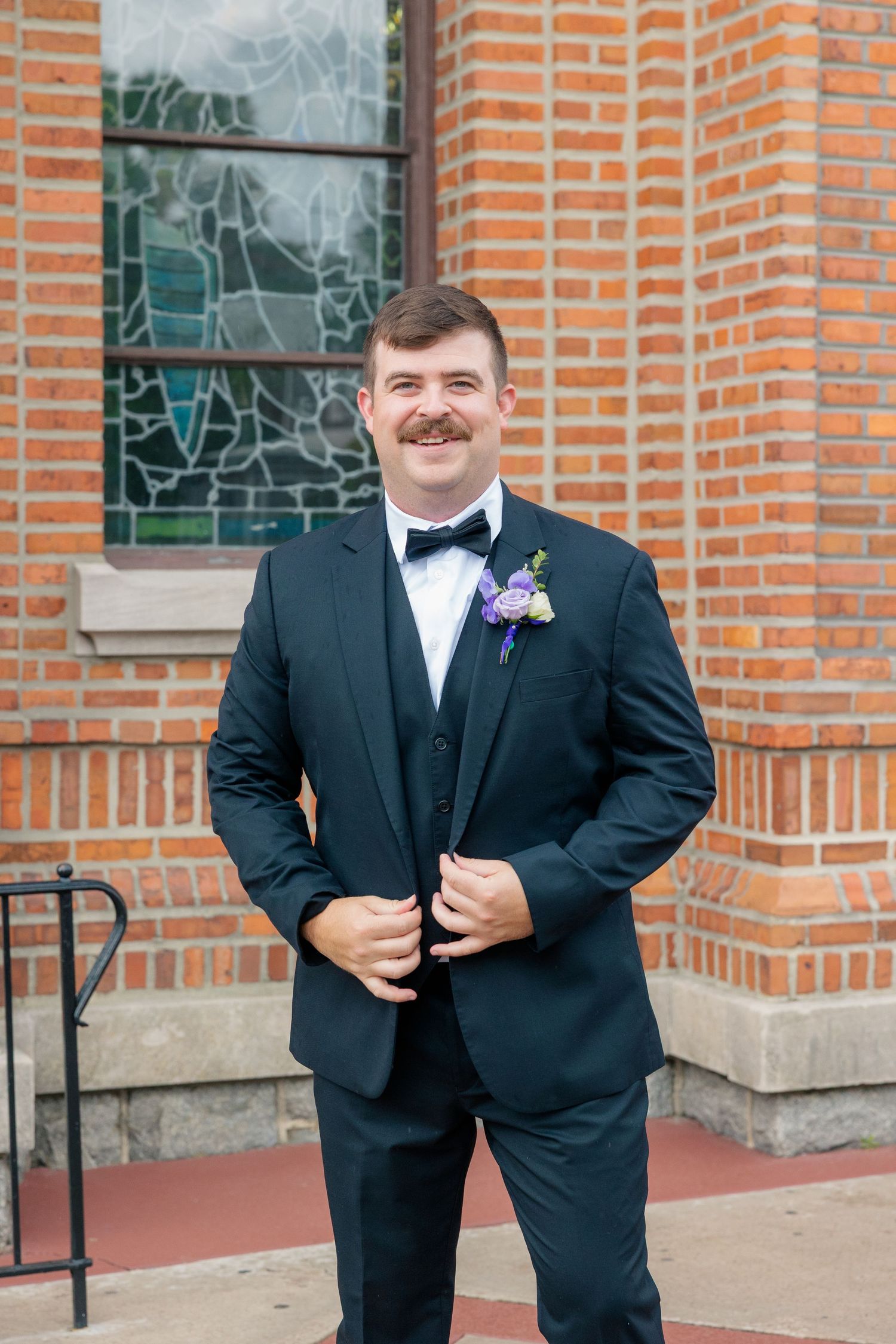 Smiling groom in black tuxedo with bow tie and purple boutonniere stands before brick church.