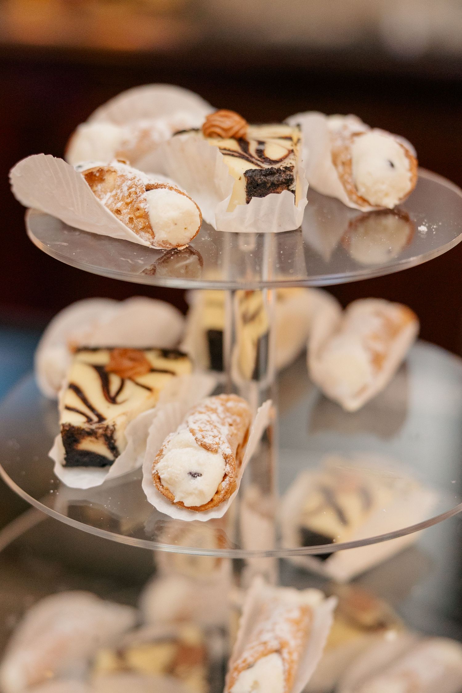 Elegant tiered glass display stand filled with assorted cannoli pastries dusted with powdered sugar at a dessert table.