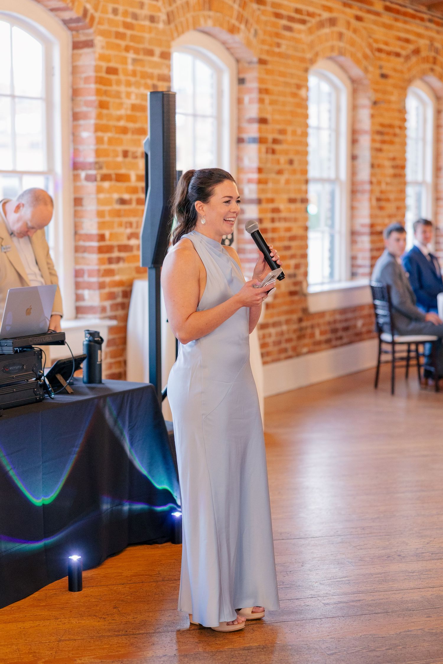 Bridesmaid in silver gown delivers heartfelt speech with microphone in elegant brick-walled wedding reception venue.