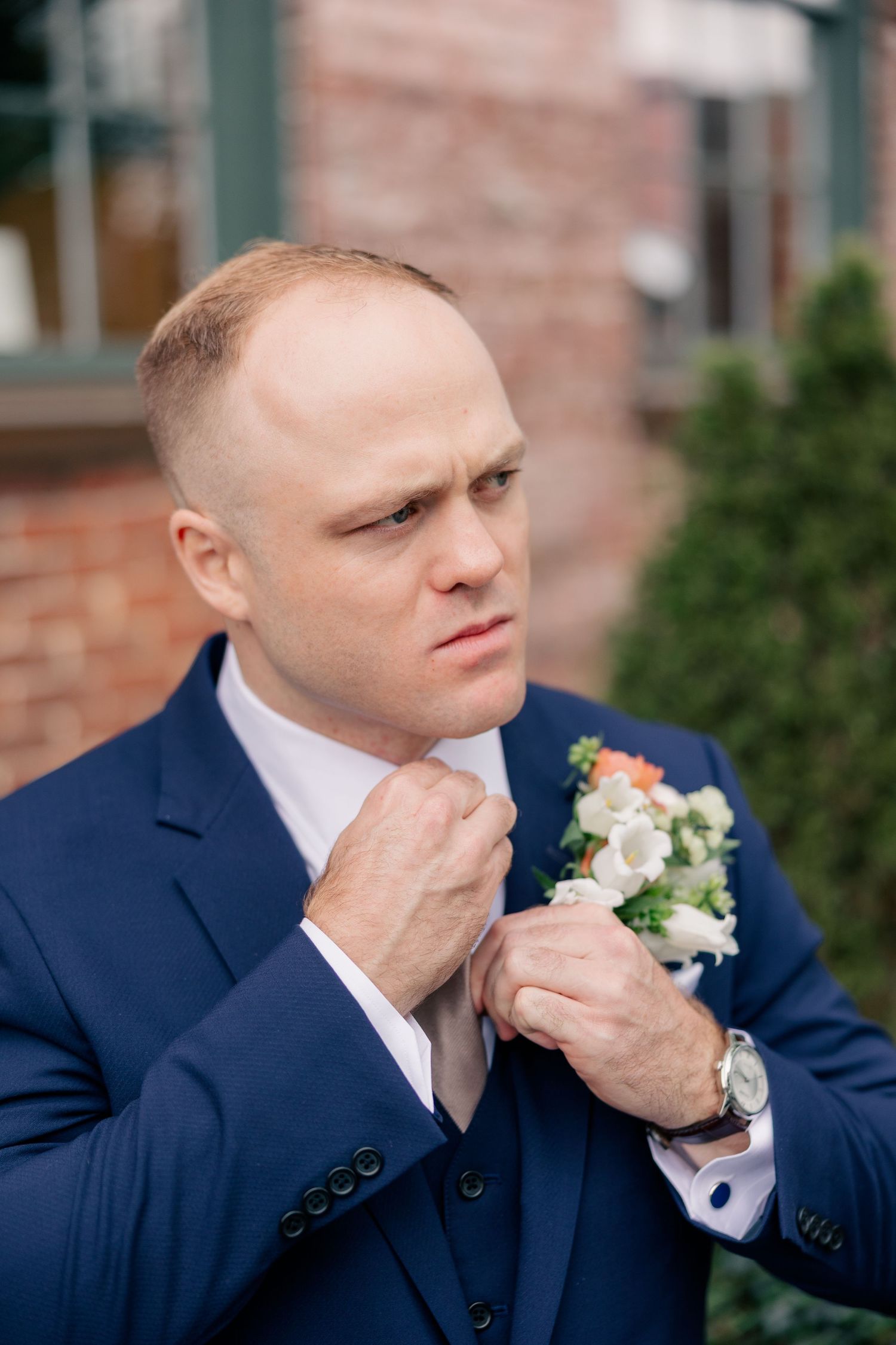 Groom in navy three-piece suit adjusts tie while pinning floral boutonniere before wedding ceremony.