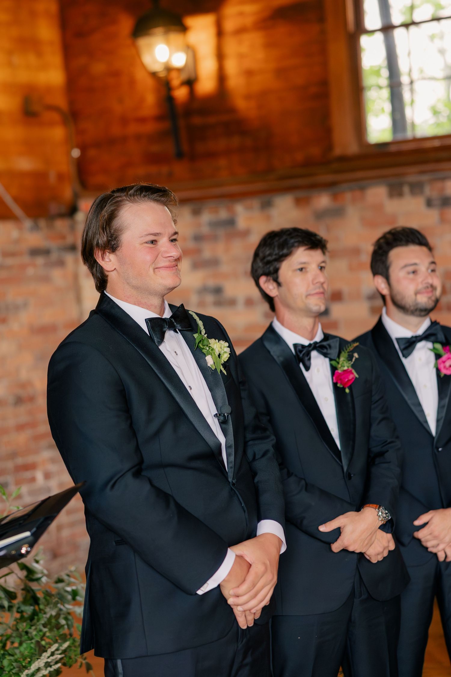 Groom smiling at altar in black tuxedo with pink boutonniere flanked by groomsmen during ceremony.