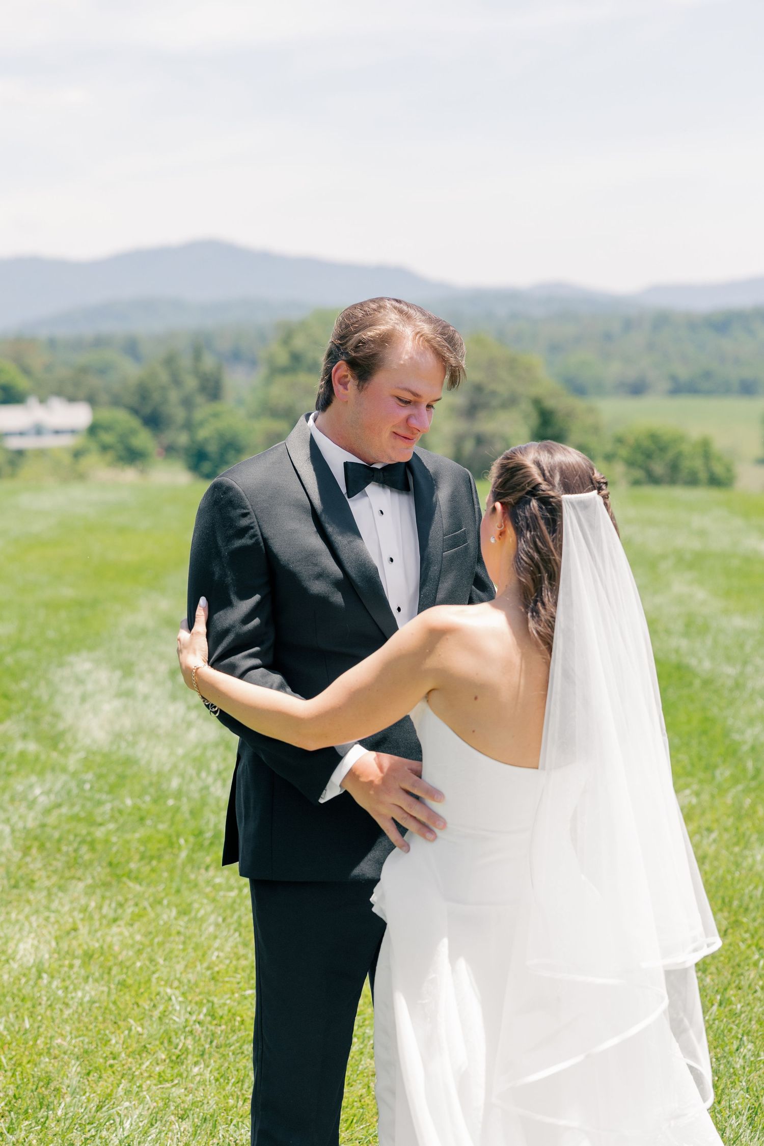 Bride in white gown with veil touches groom in black tuxedo during first look with mountain backdrop.