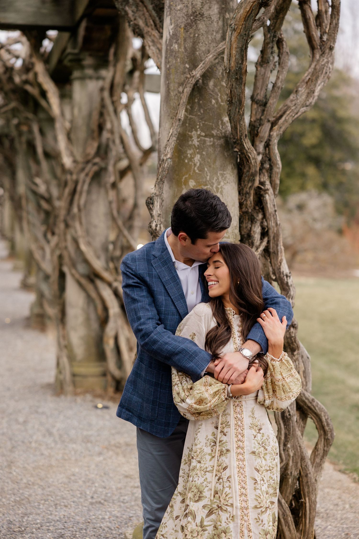 A couple embraces lovingly under twisted tree branches on a garden path.
