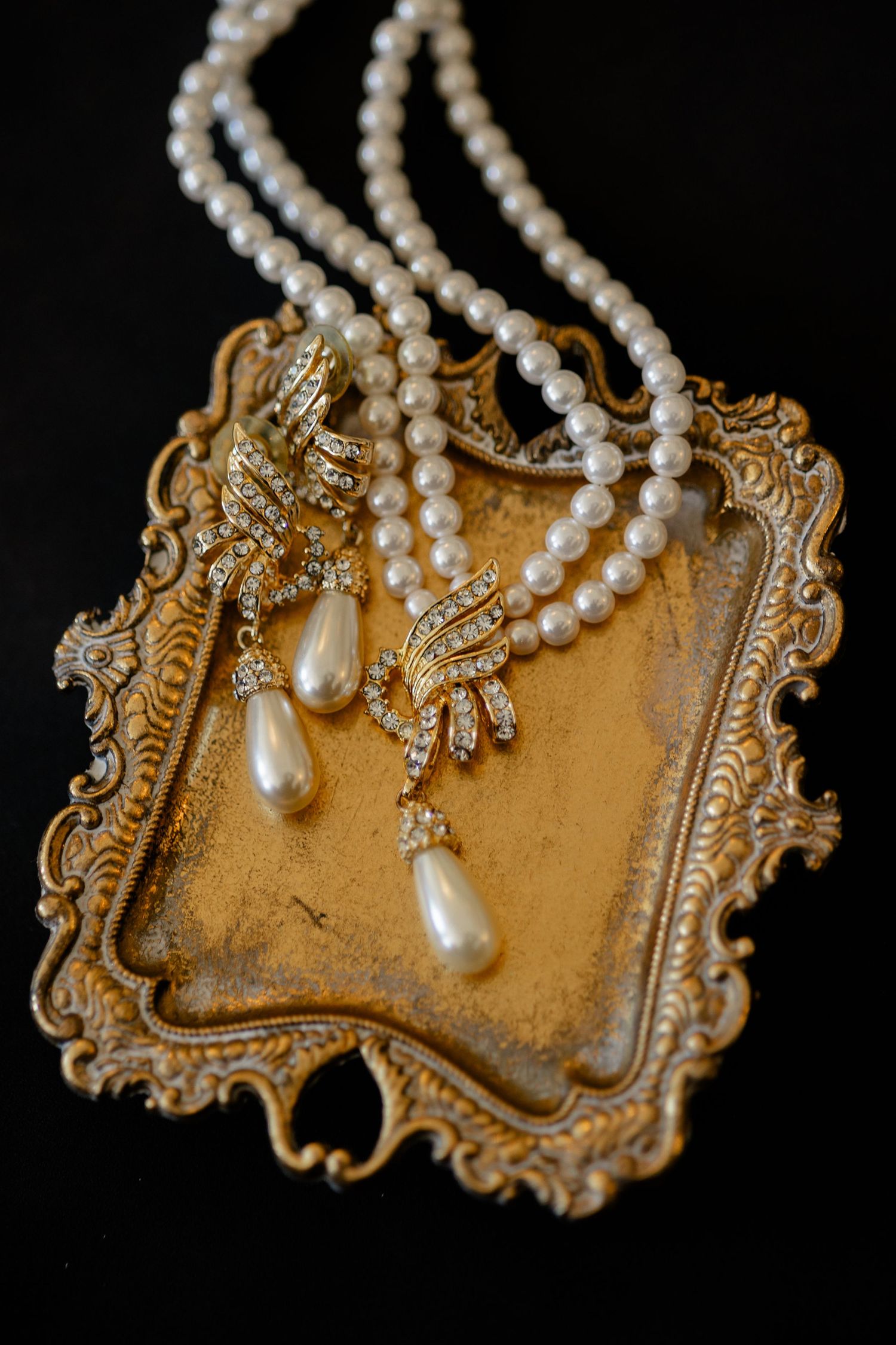 Elegant pearl necklace and teardrop jewelry displayed on ornate vintage gold frame against black background.