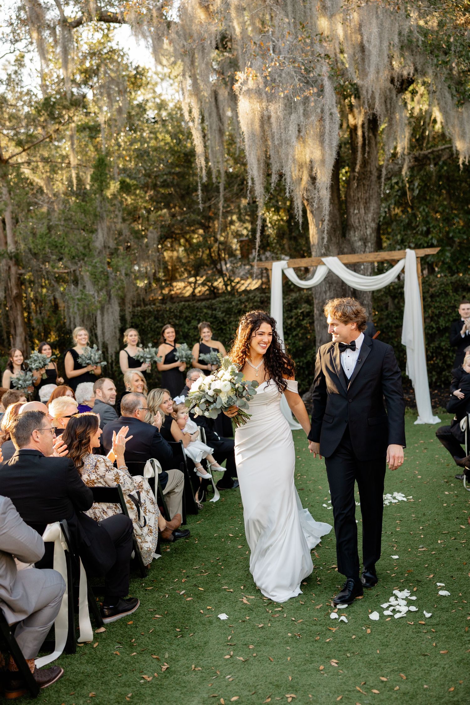 A wedding ceremony under Spanish moss-draped trees with guests seated on either side of a white-draped arch.