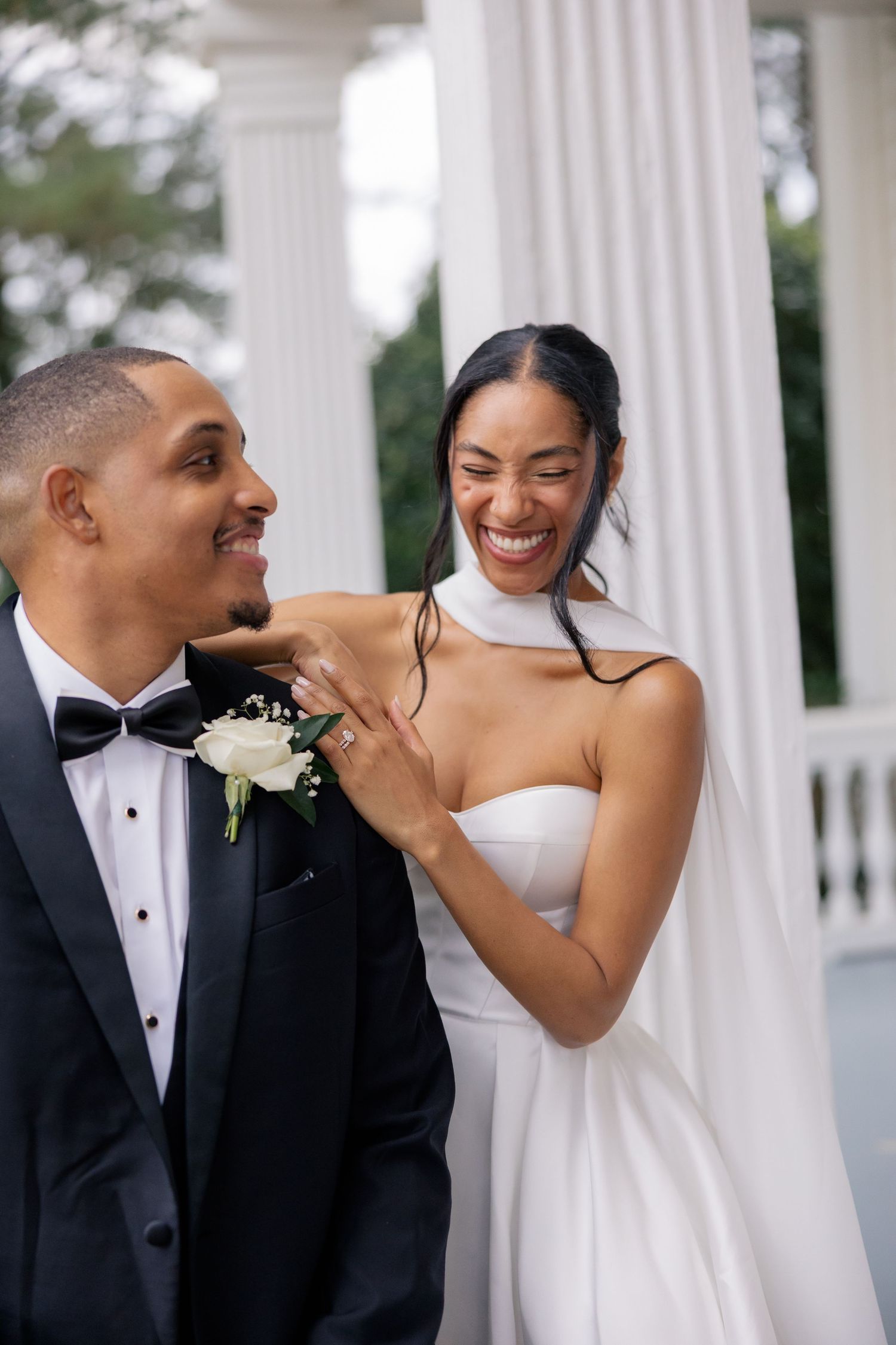 A joyful bride in a white strapless gown laughs beside her groom in a black tuxedo with boutonniere outdoors.