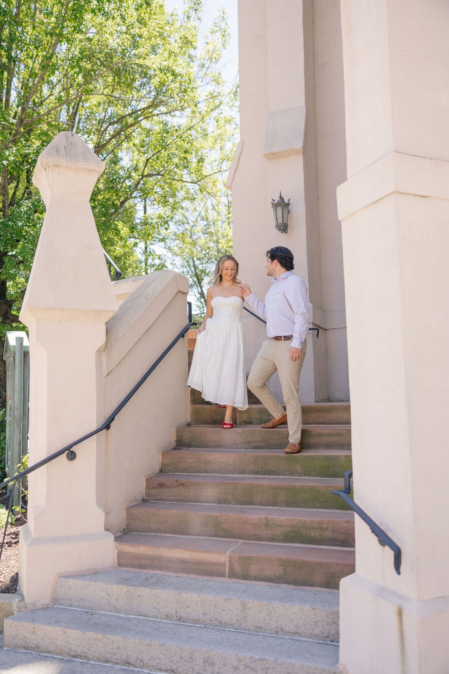 Couple holding hands walking down elegant stone staircase between white columns surrounded by trees.