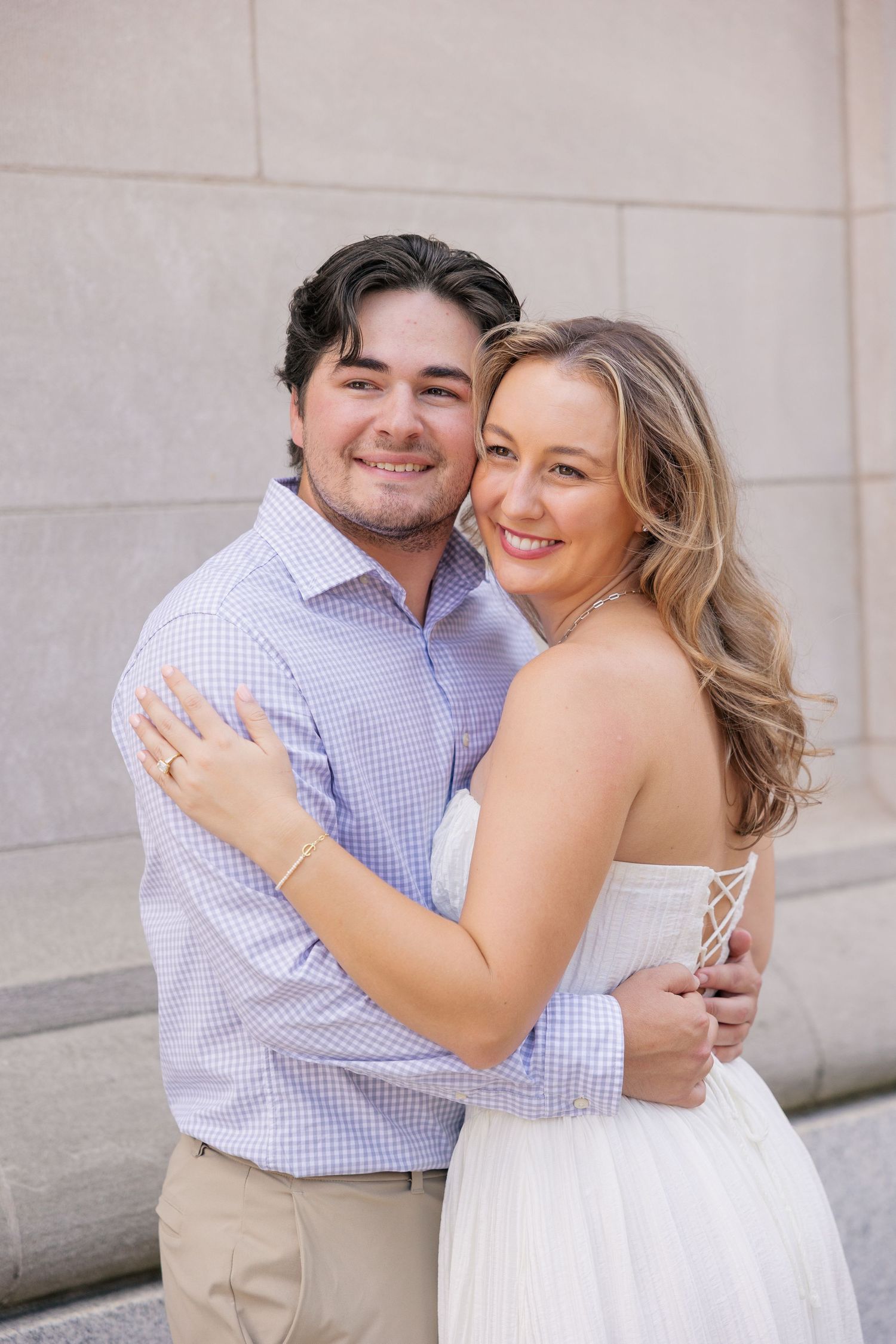 Smiling couple embracing near stone columns, woman in white dress and man in blue gingham shirt, engagement photo.