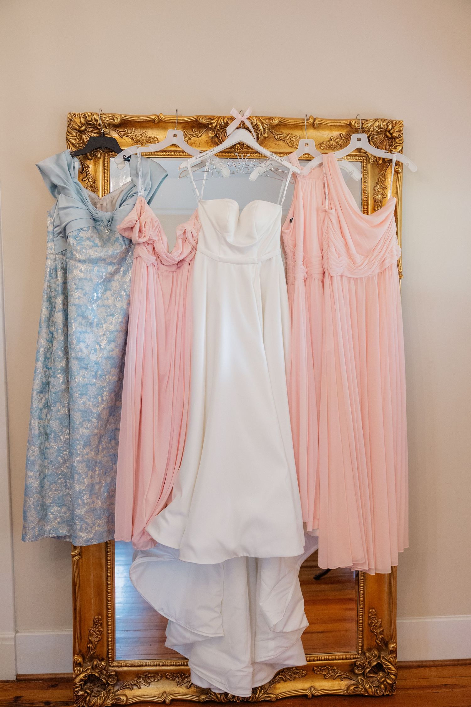 Pastel pink, white, and blue dresses hanging against an ornate gold-framed mirror in a bridal boutique.