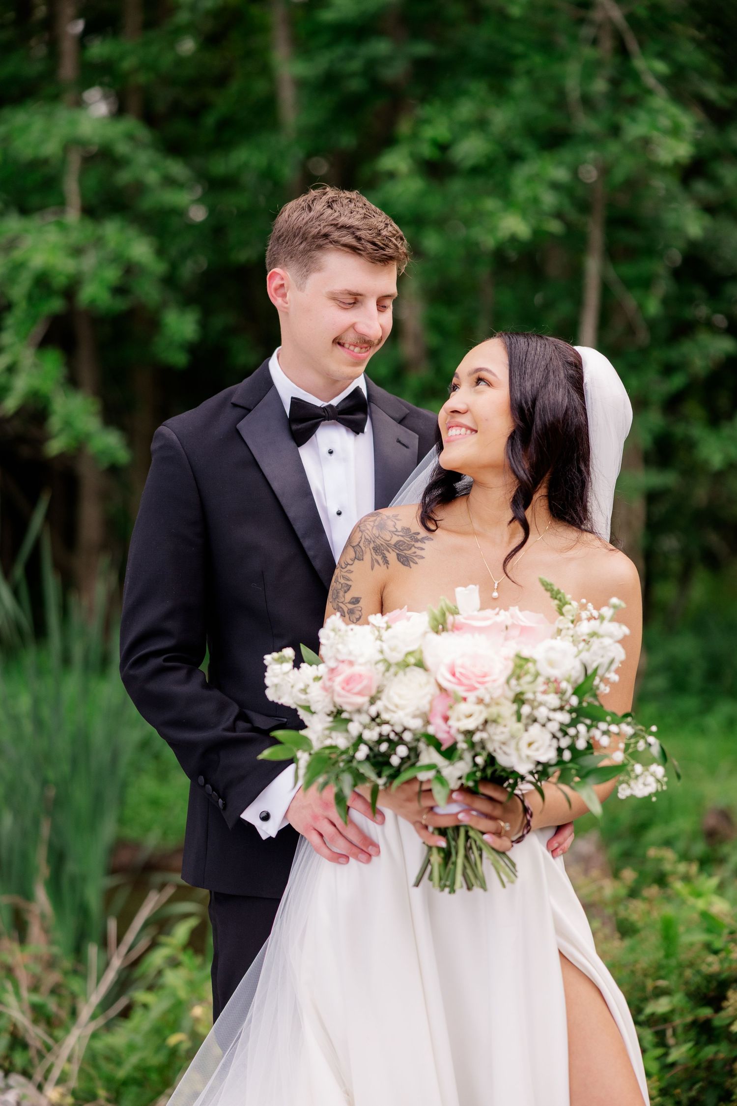 Newlyweds share a joyful moment outdoors while holding pink and white bouquets.