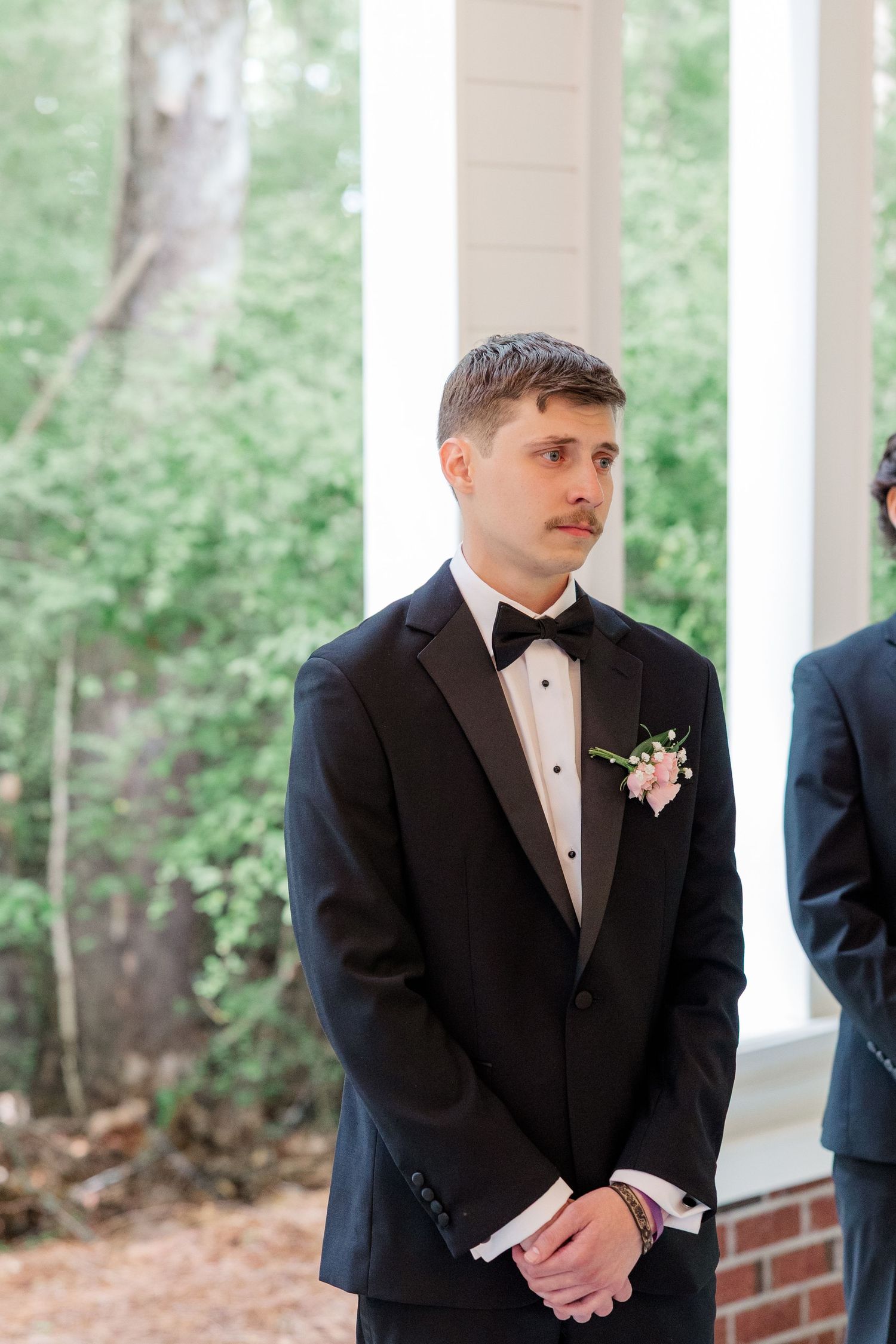 Groom in classic black tuxedo with boutonniere standing at outdoor wedding ceremony venue.